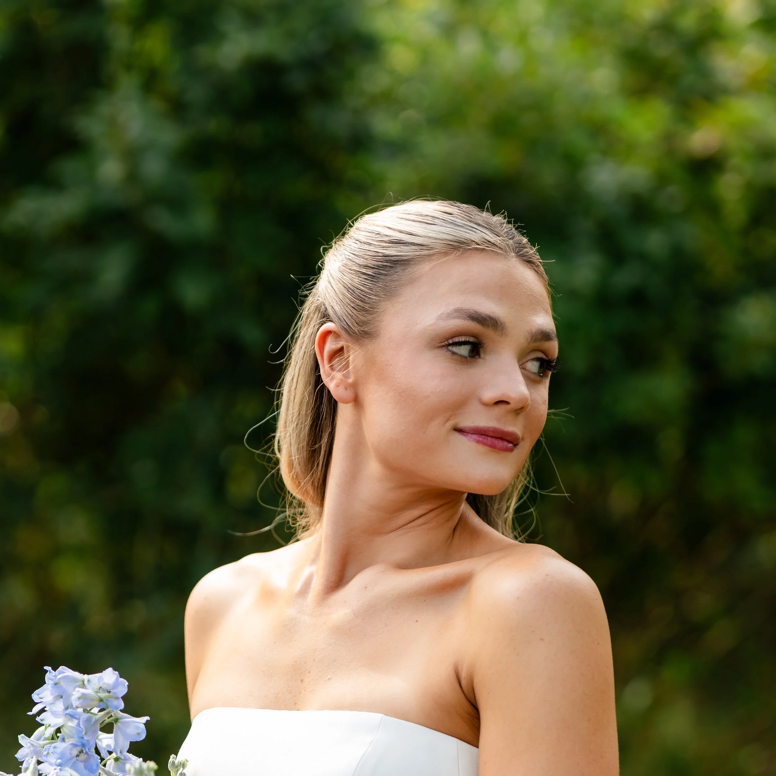 A young woman with blonde hair styled back, looking to the right with a slight smile, standing outdoors with green trees in the background, wearing a white strapless dress.