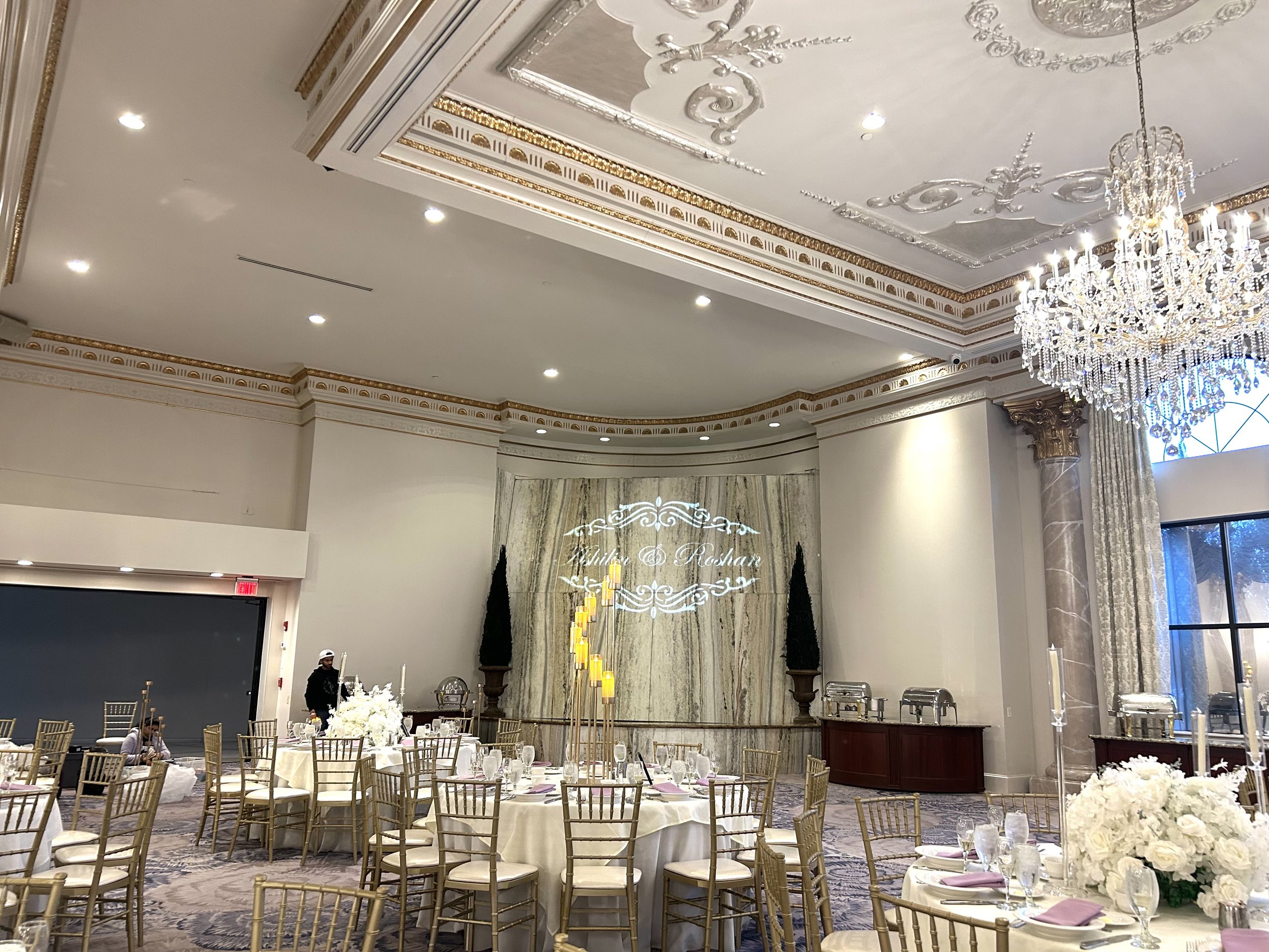 Elegant banquet hall with round tables covered in white tablecloths, set with glassware and pink napkins, gold chairs, a grand chandelier, and a decorative wall with floral arrangements and projected names, for a wedding celebration.