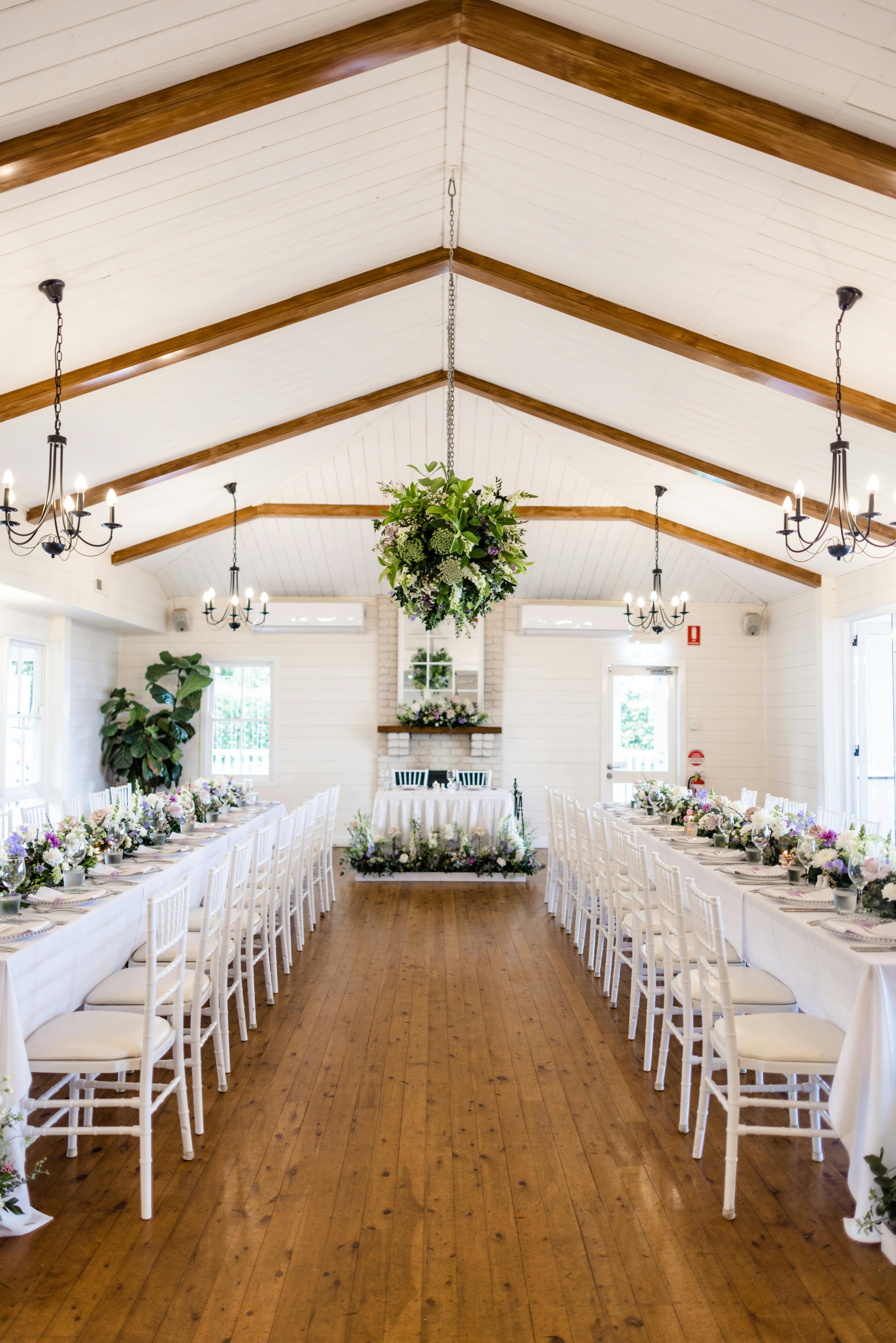 Interior of a wedding reception hall with white walls and wooden floor, decorated with floral centerpieces on long tables, chandeliers hanging from the ceiling, and a floral chandelier as a centerpiece above the aisle leading to an altar.