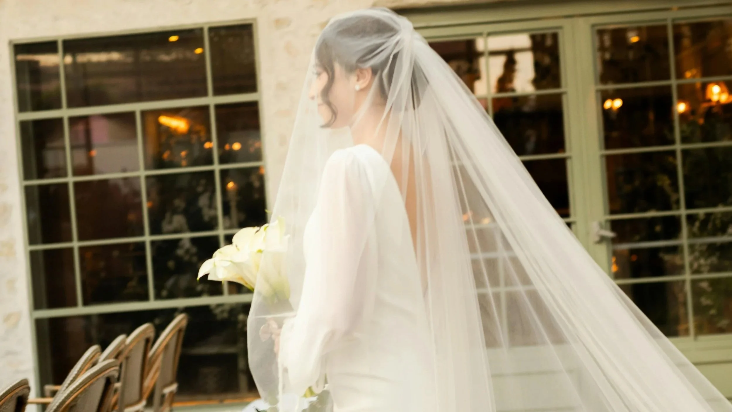 A bride in a white wedding dress holding a bouquet of white calla lilies, wearing a long veil, standing indoors with large window panes in the background.
