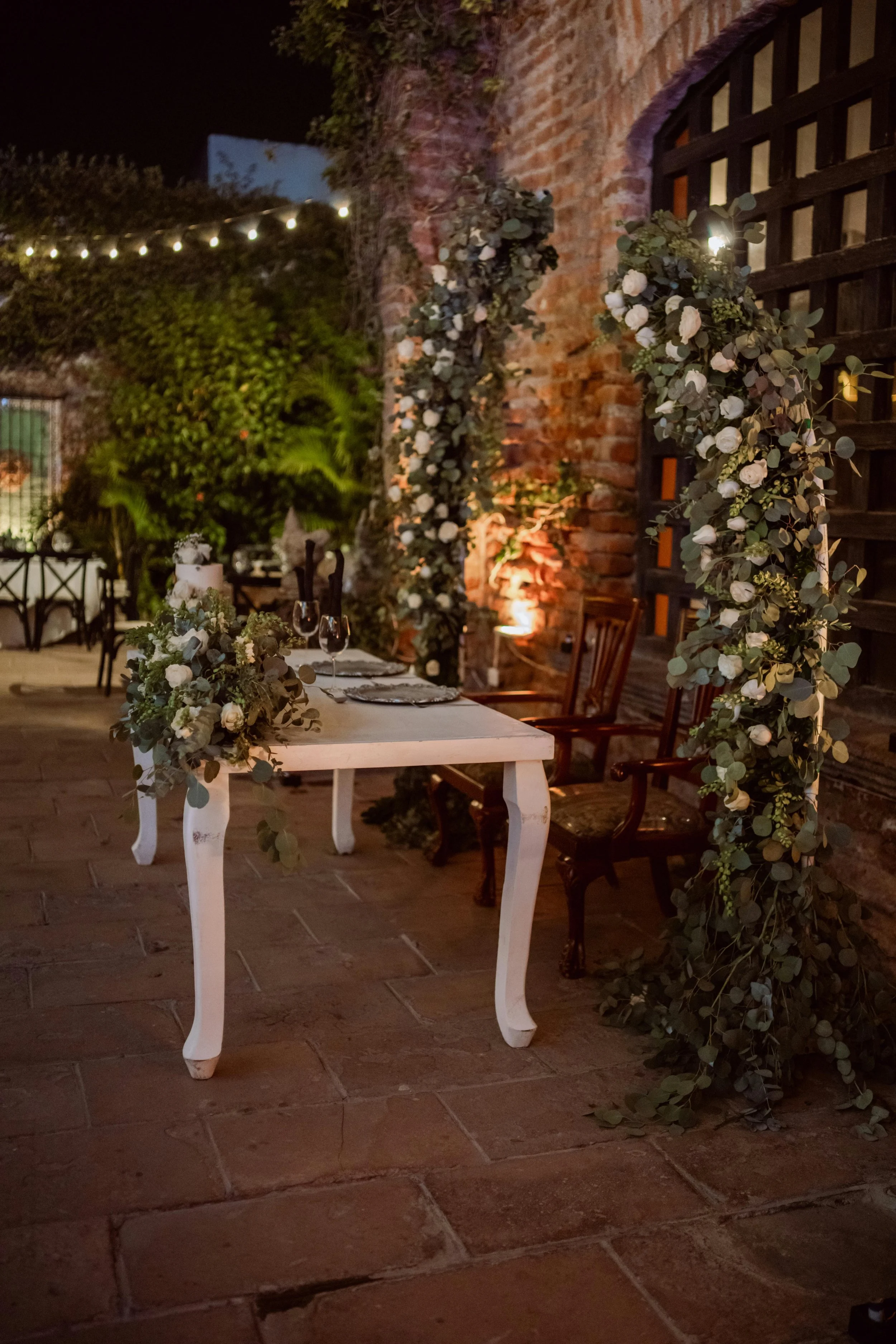 An outdoor decorated table setting at night with a white table, floral arrangements, two glasses, and a wooden chair, surrounded by greenery and brick walls.
