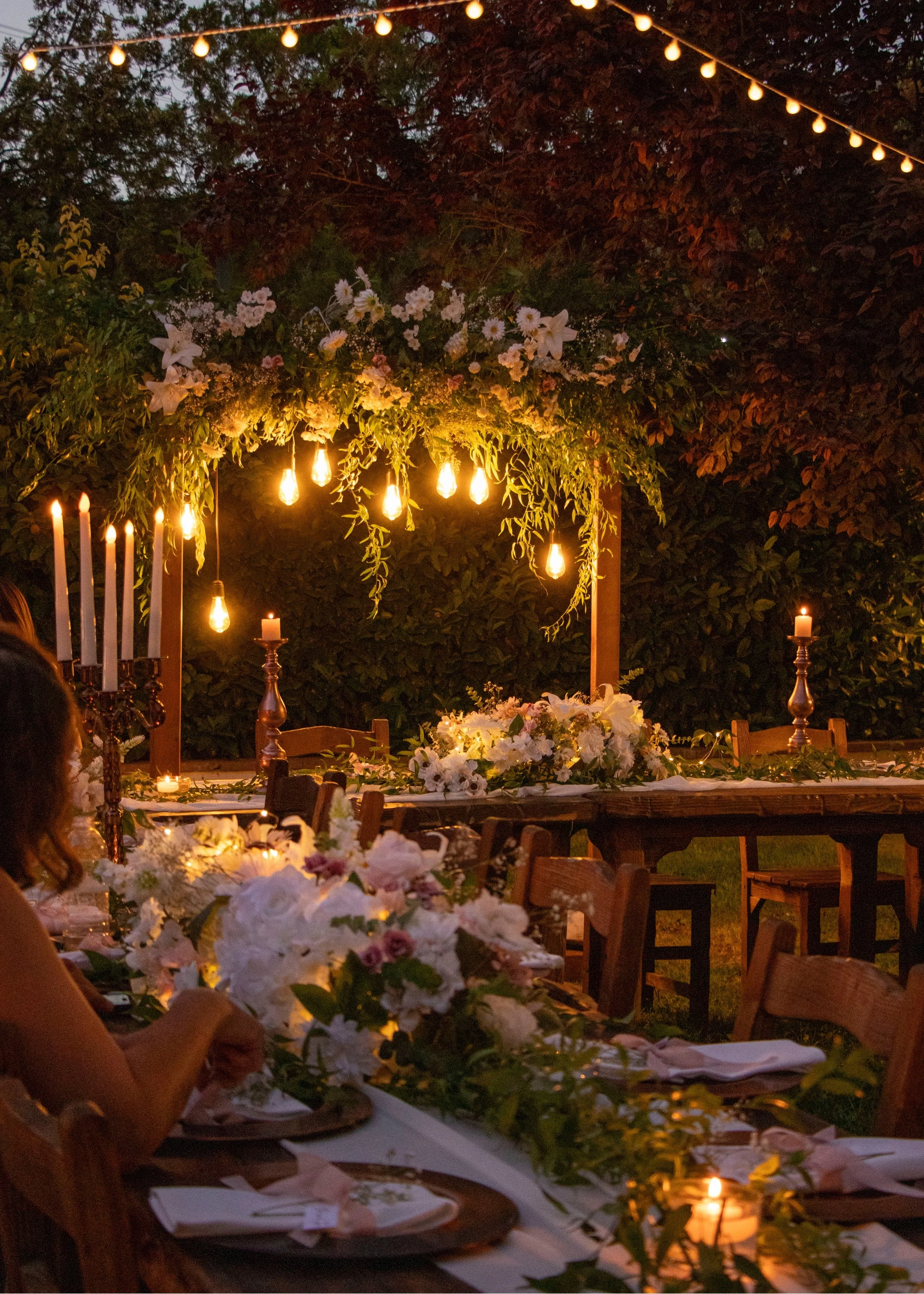 An outdoor evening dinner setup with a long table decorated with white floral arrangements, candles, and hanging Edison bulbs, surrounded by trees and foliage.