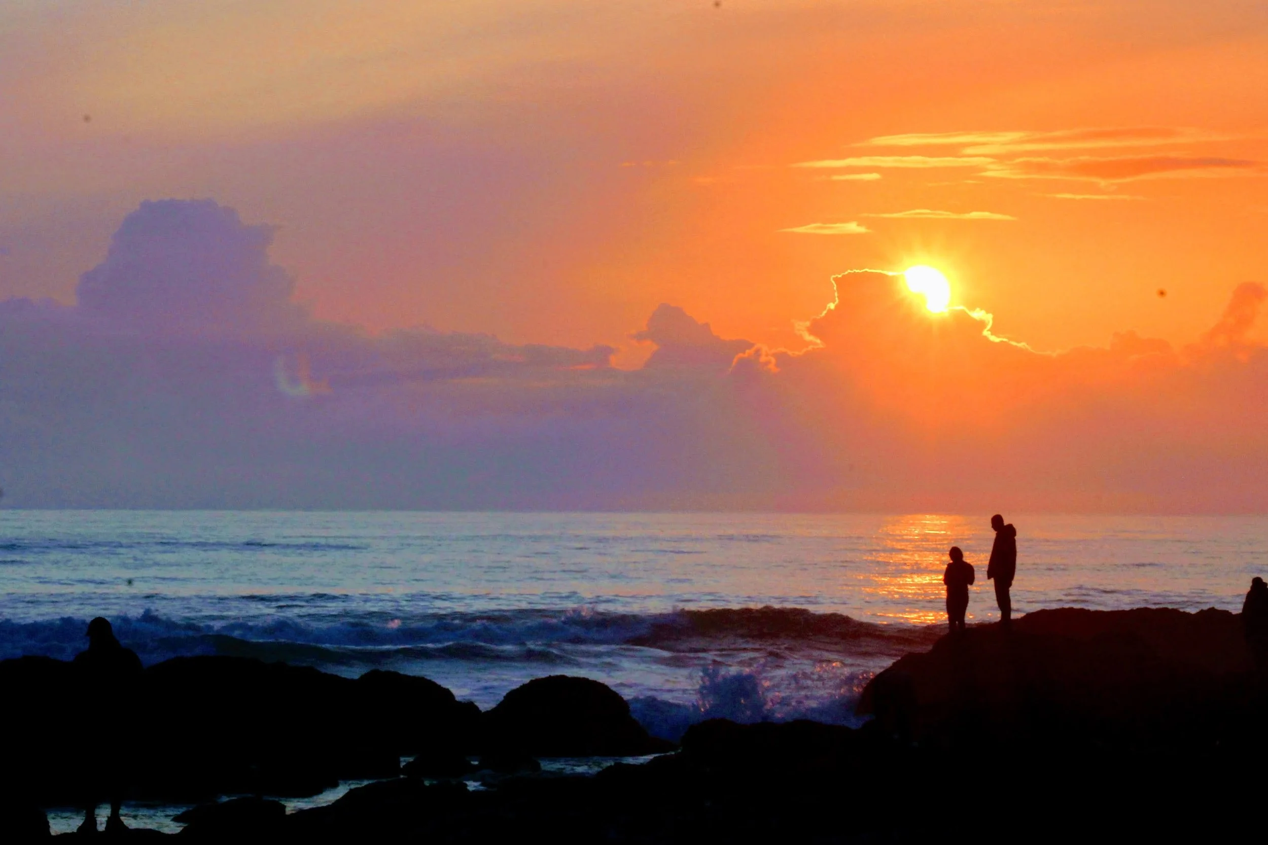 Silhouettes of two people standing on rocks by the ocean at sunset, with a vibrant orange and purple sky and the sun partially hidden behind clouds.