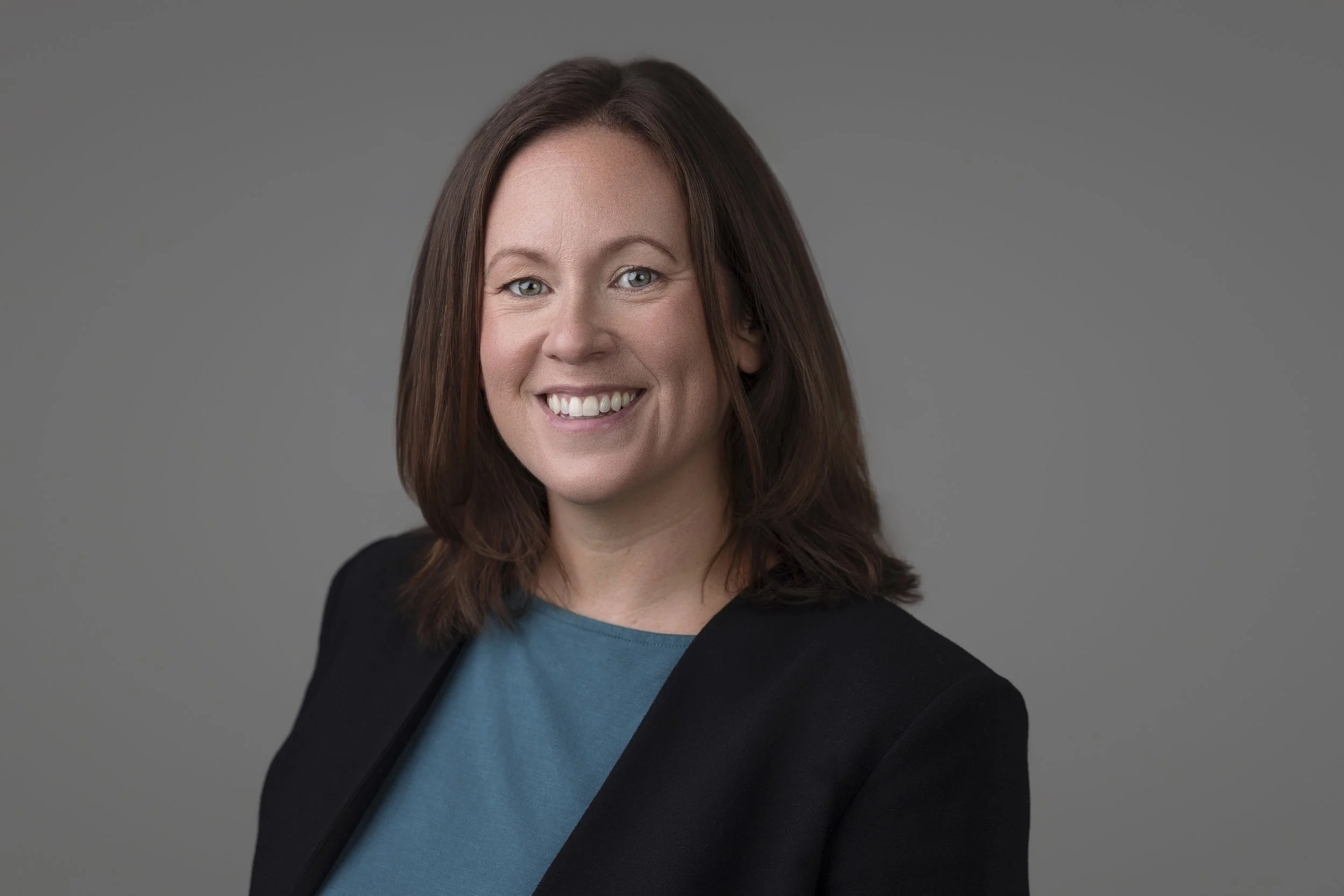 A woman with shoulder-length brown hair, wearing a blue top and black blazer, smiling against a grey background.