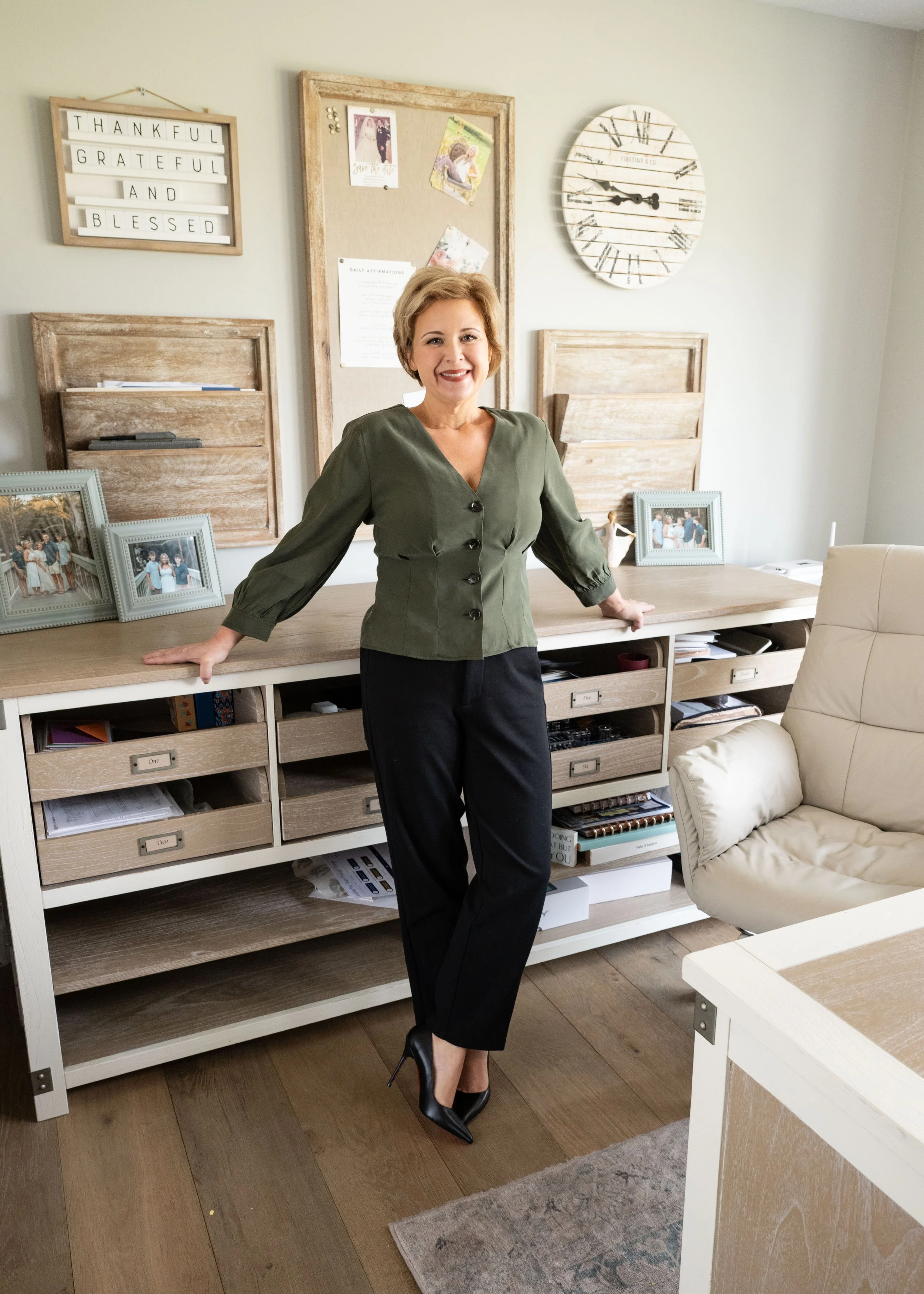 Woman in a green blouse and black pants standing in an office space, smiling, with a light-colored wall, wooden and white furniture, framed photos, and decorative items in the background.