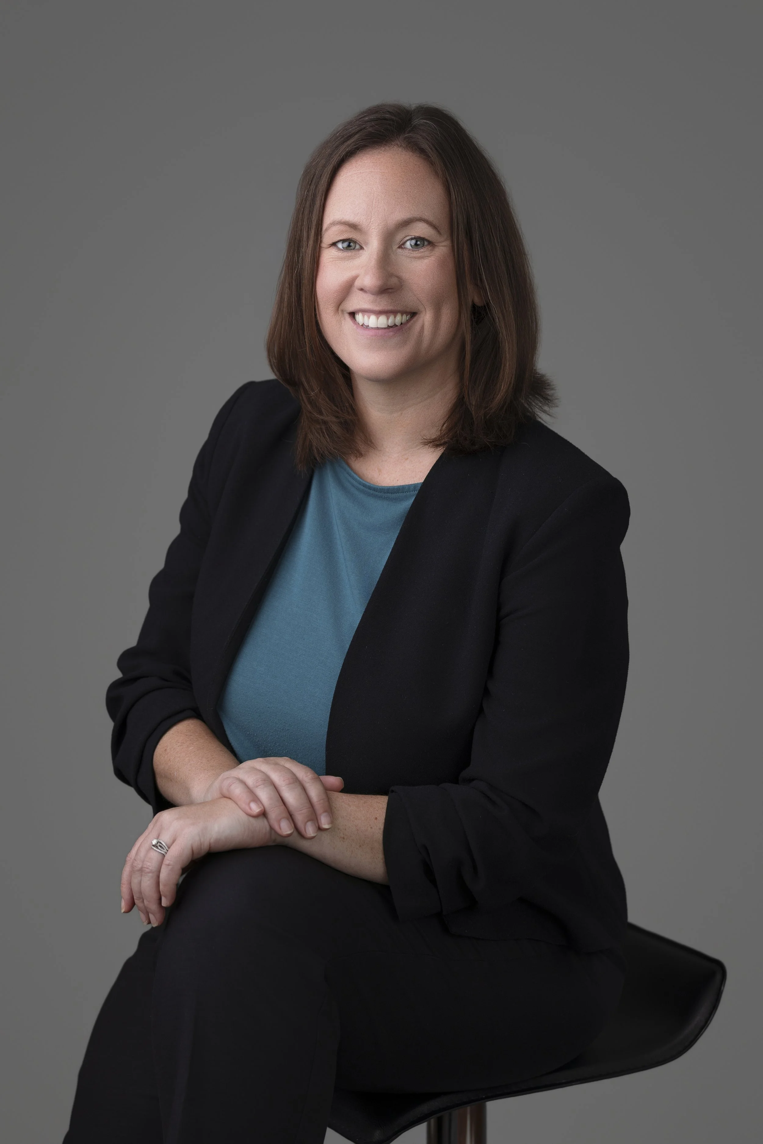 A woman with shoulder-length brown hair, smiling, wearing a black blazer over a teal top, sitting on a black chair against a gray background.