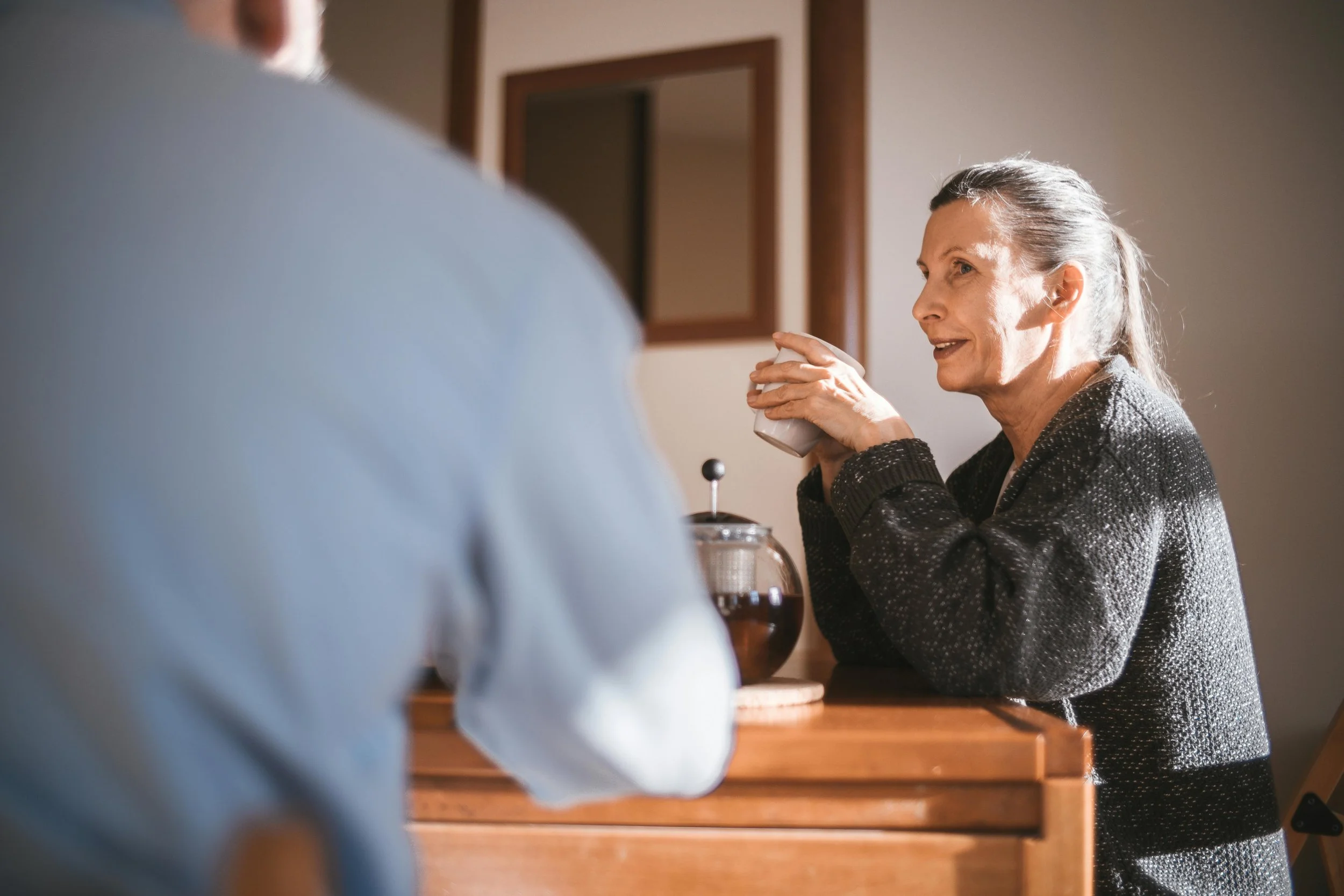 An elderly woman with gray hair smiling and talking to a person across from her, holding a coffee mug, sitting at a wooden table in a cozy home setting.