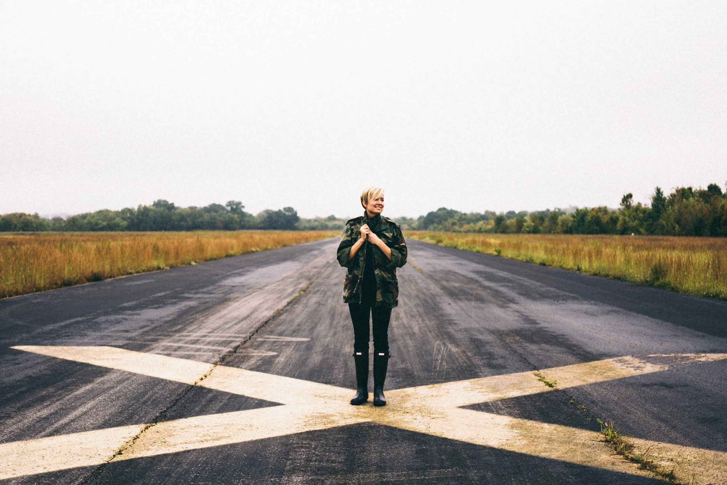 A woman standing in the middle of an empty road with large white X markings, surrounded by open fields and trees under a cloudy sky.