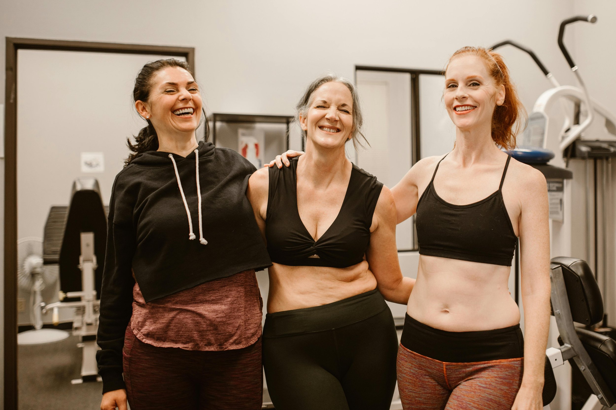 Three women in workout clothes smiling and embracing in a gym.