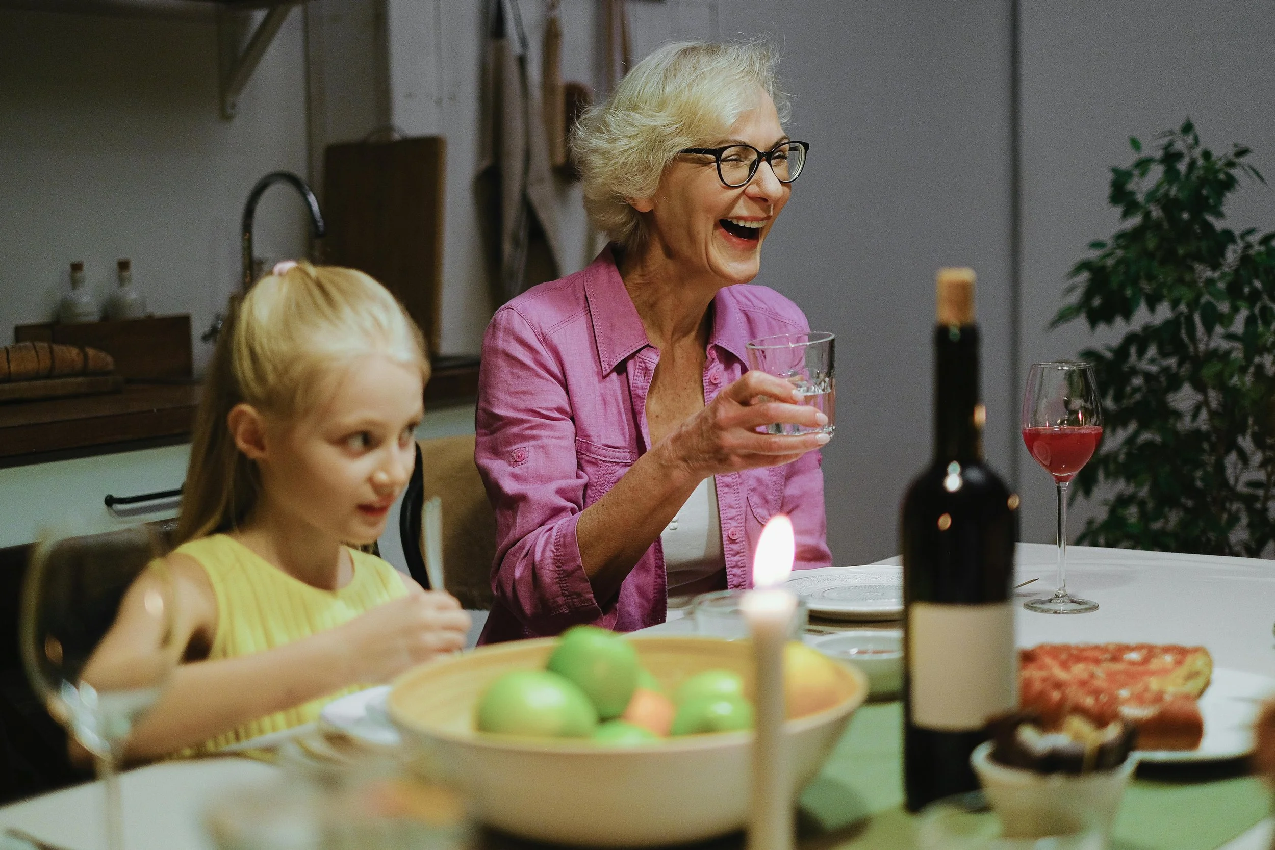 An elderly woman laughing and holding a glass of water sitting at a dinner table with a young girl, surrounded by food and drinks.