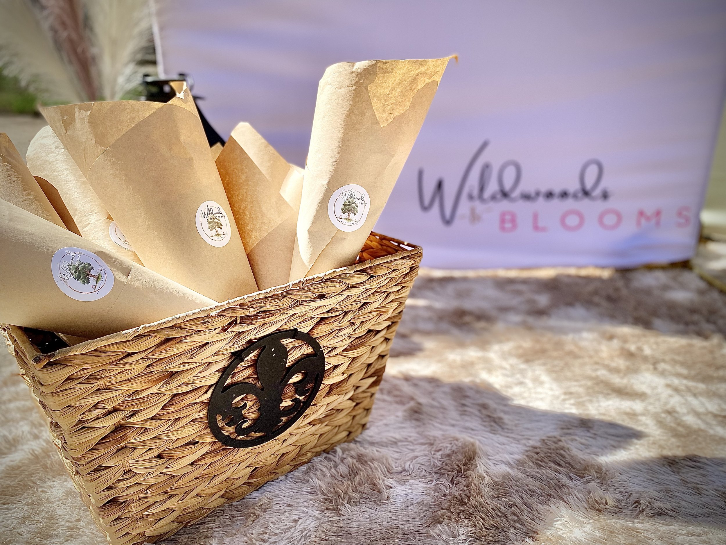 A wicker basket with a black octopus logo contains beige paper cones with stickers, placed on a plush beige surface with a background showing a sign that reads "Wallwoods BLOOMS".