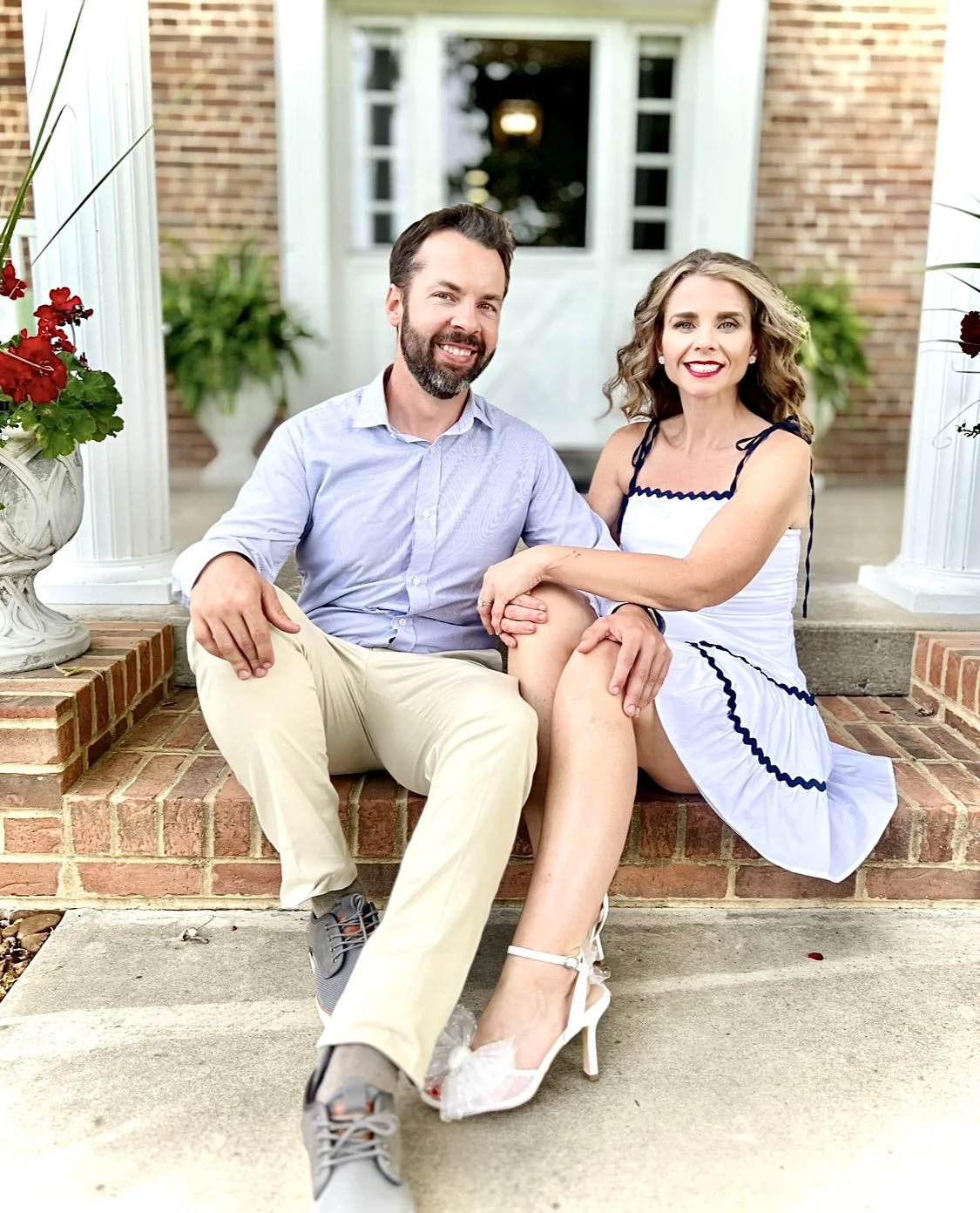 A smiling couple sitting on the steps of a brick porch, posed closely together with greenery and white columns in the background.