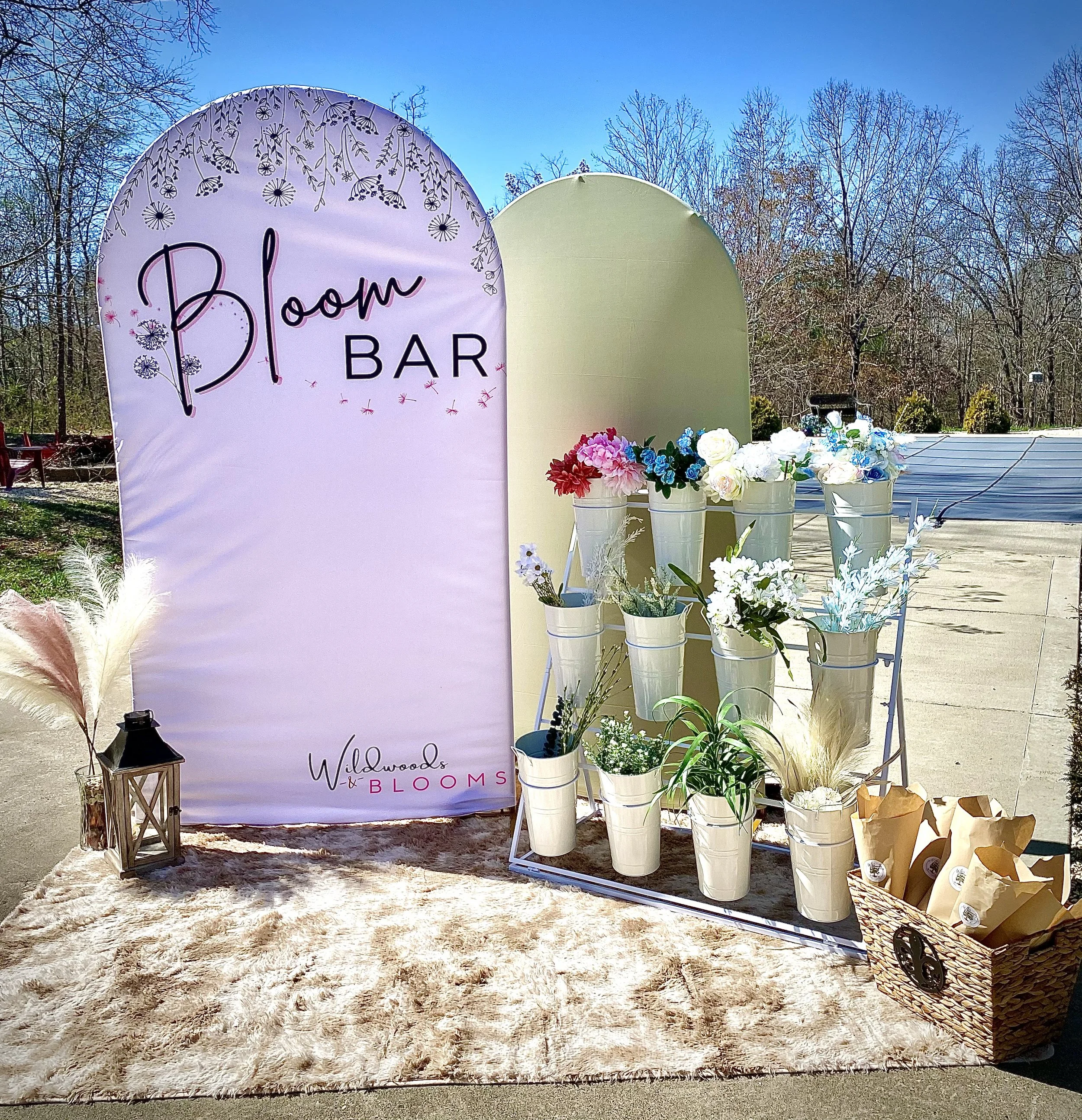 A floral display setup for a Bloom Bar at an outdoor event. It includes a large sign with the words 'Bloom Bar' and a floral design, white buckets filled with flowers arranged on a stand, a fur rug, a lantern, and a basket with wrapped gifts or favors. The background shows trees and a clear blue sky.