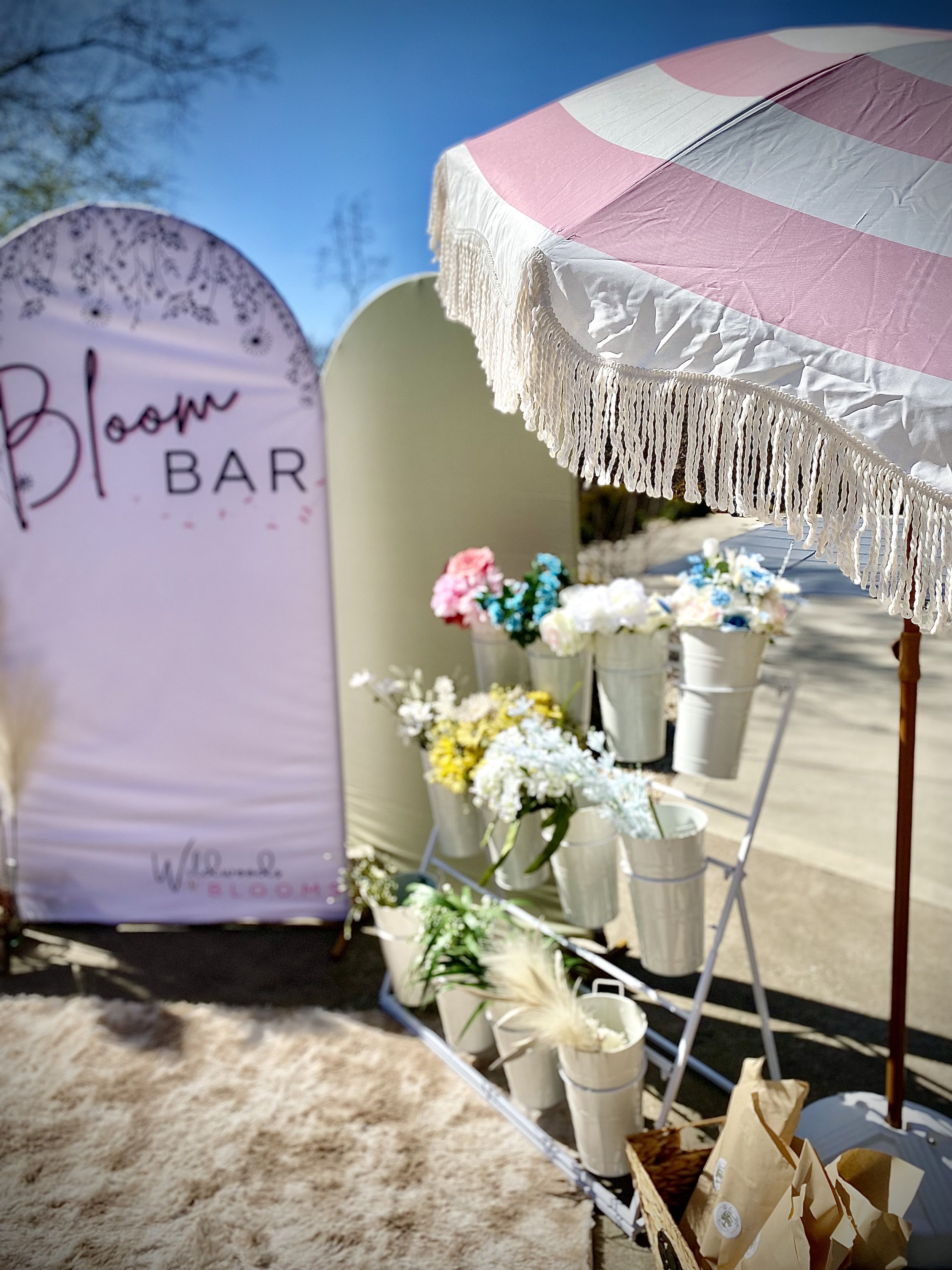 Bloom bar flower stand with white buckets filled with colorful local flowers, beneath a pink and white striped parasol, with a pink sign that reads 'Bloom Bar' in the background on a sunny day.