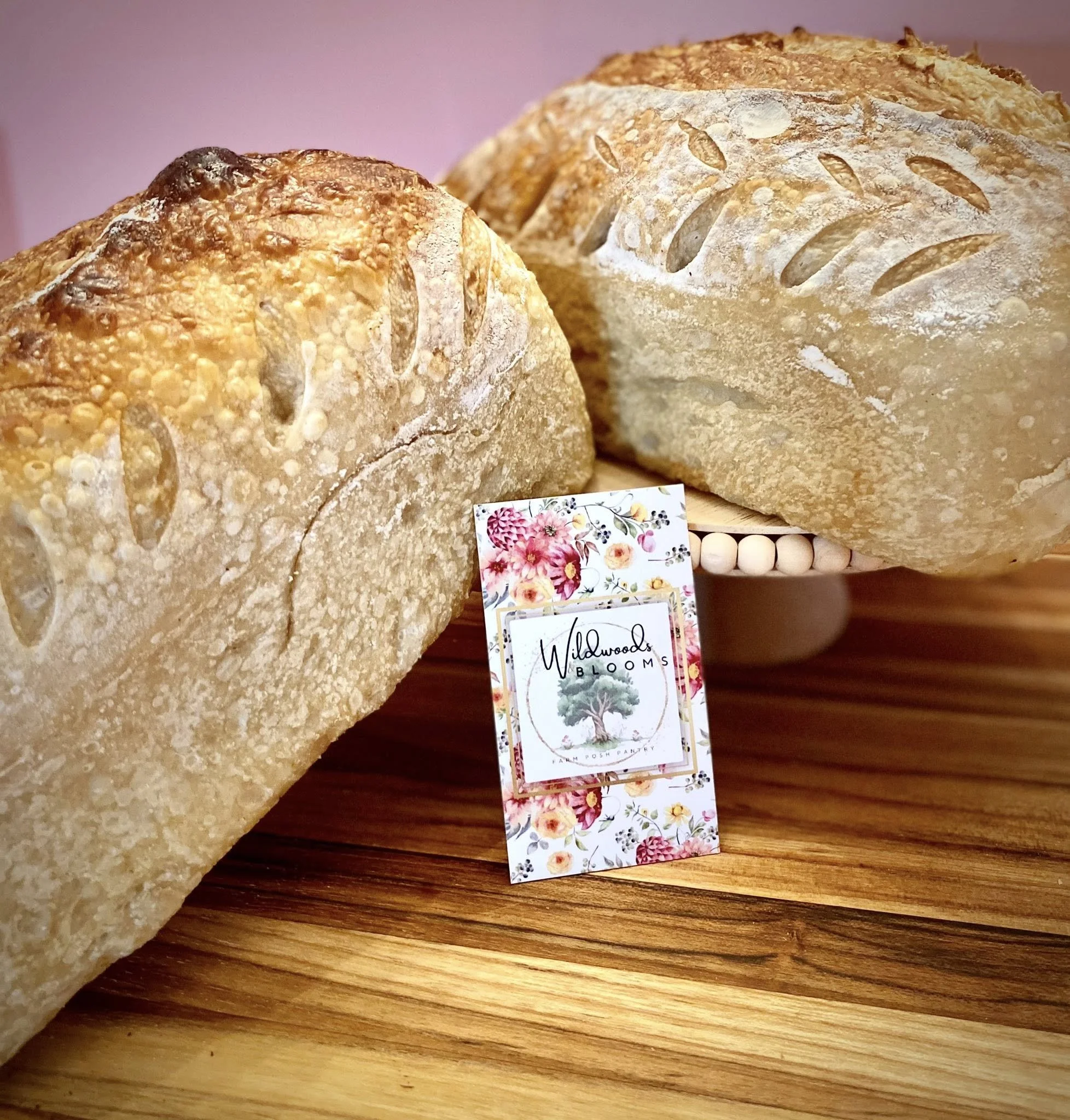 Close-up of two loaves of rustic sourdough bread with a floral business card that reads 'Wildwoods Blooms' Cadiz KY, placed on a wooden surface.