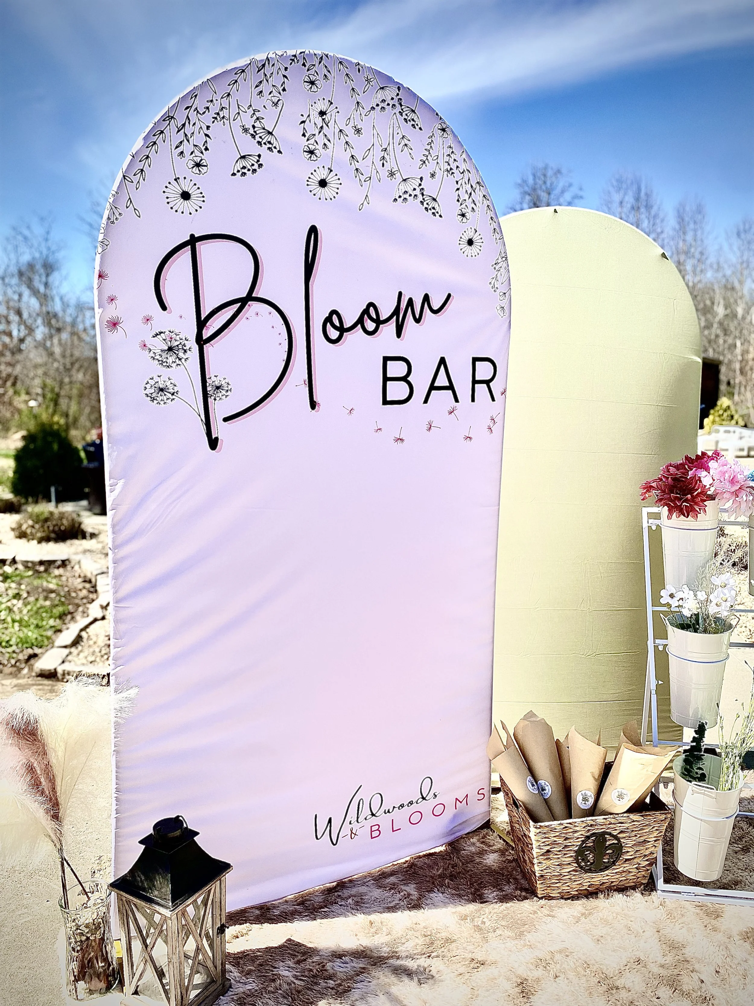 A light purple, arched sign with black and pink lettering that says 'Bloom Bar,' decorated with floral illustrations at the top. To the right, there are white containers with pink and white flowers, and to the left, a small black lantern and a basket with wrapped bouquets of flowers. The scene is outdoors on a sunny day.