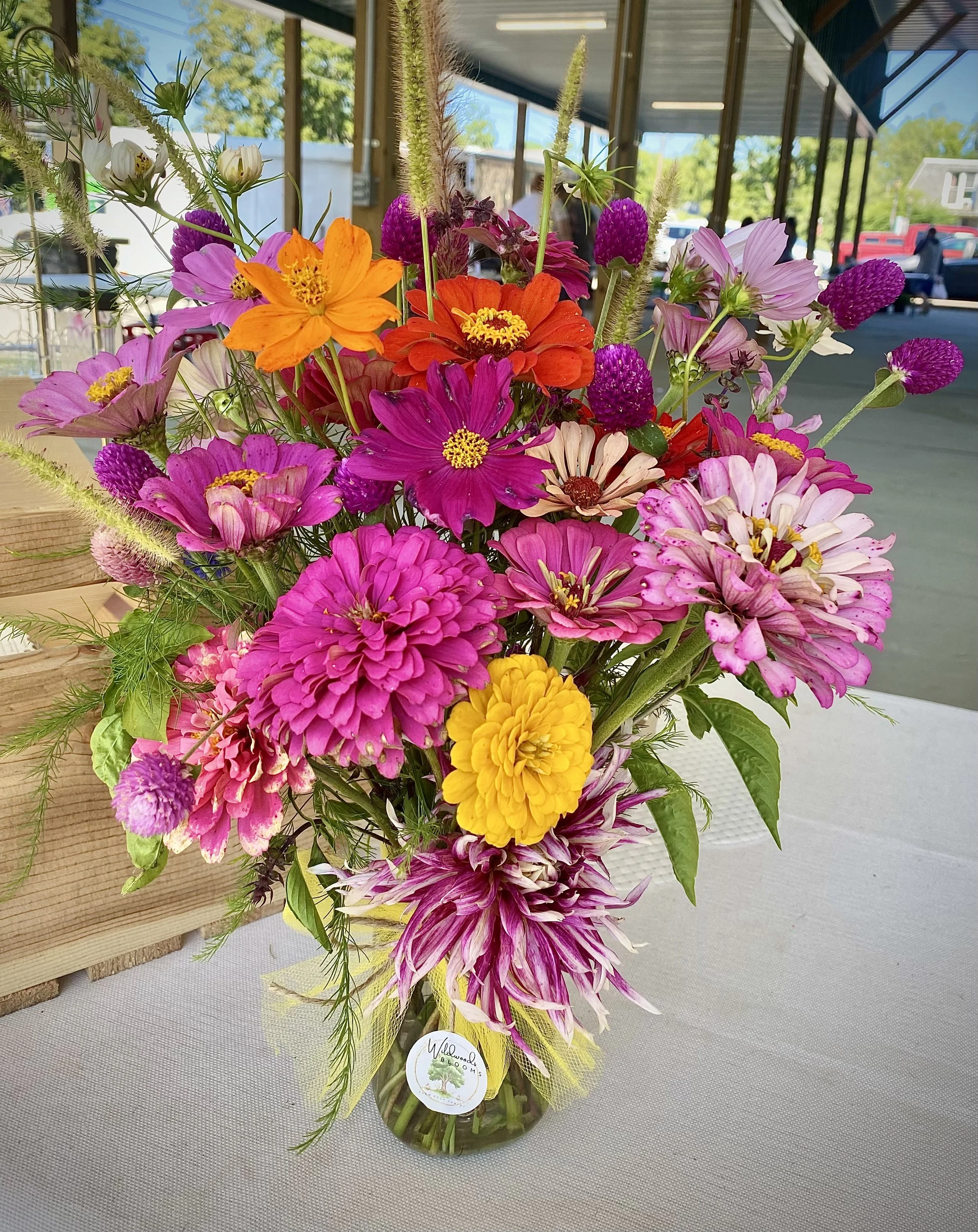 A colorful CSA bouquet of mixed flowers in a glass vase on a table outside, with a building, cars, and green trees in the background.