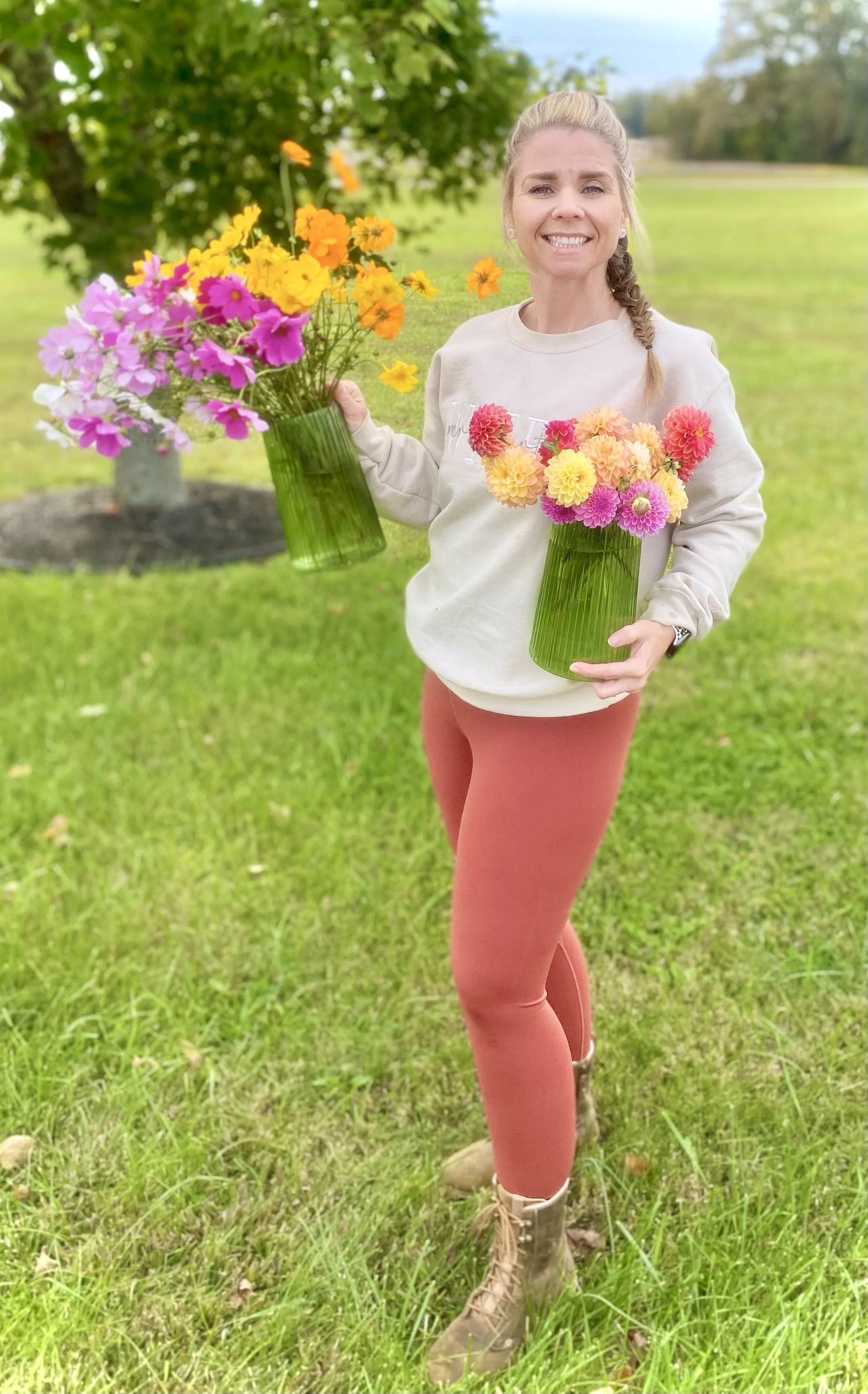 A woman standing outdoors on grass, holding two green vases with colorful flowers, smiling at the camera.
