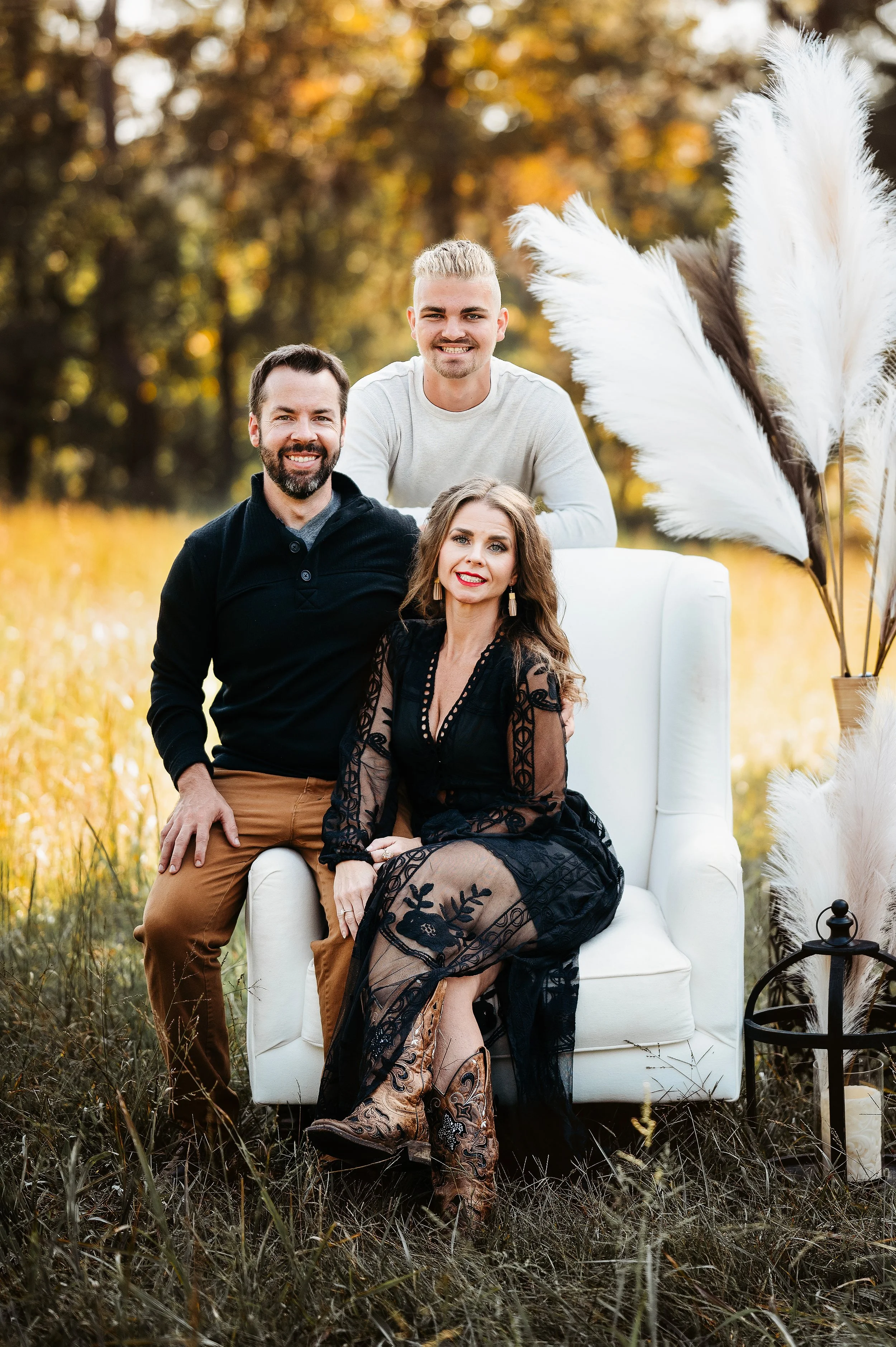 A family outdoors in a field with autumn foliage, sitting on and around a white armchair. They are smiling and dressed in casual and stylish clothing. Tall white pampas grass decorates the scene, and there is a black lantern on the ground nearby.