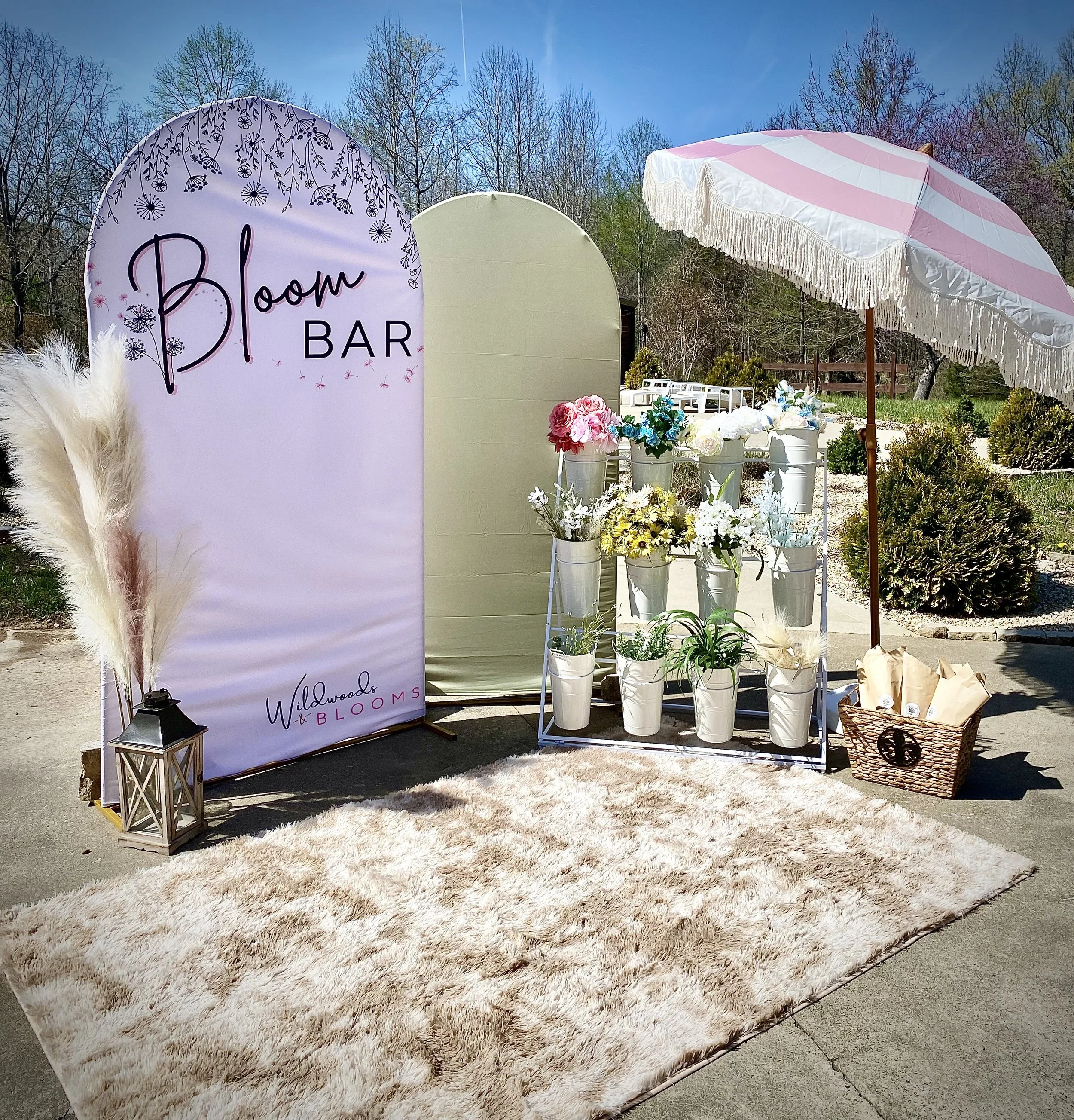 Outdoor floral display with a pink and white striped umbrella, white bucket flower arrangements, a pink sign reading 'Bloom Bar,' and a beige rug on concrete, set against a background of trees and blue sky.