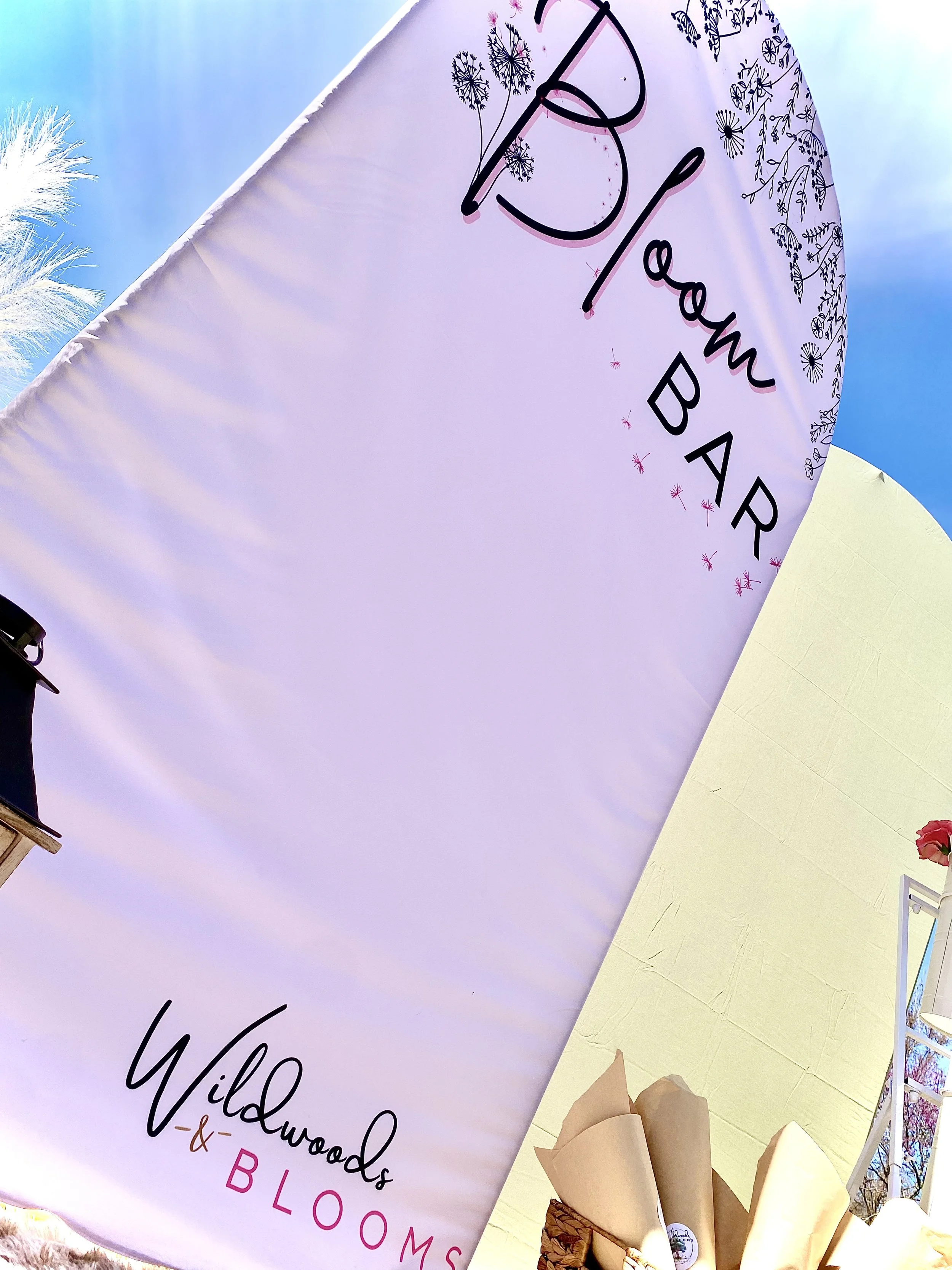 A large vertical banner with the text 'Bloom Barn' at the top, decorated with floral illustrations. The bottom part of the banner reads 'Wildwoods & BLOOMS'. The scene appears to be outdoors, with a blue sky and some plants visible around.