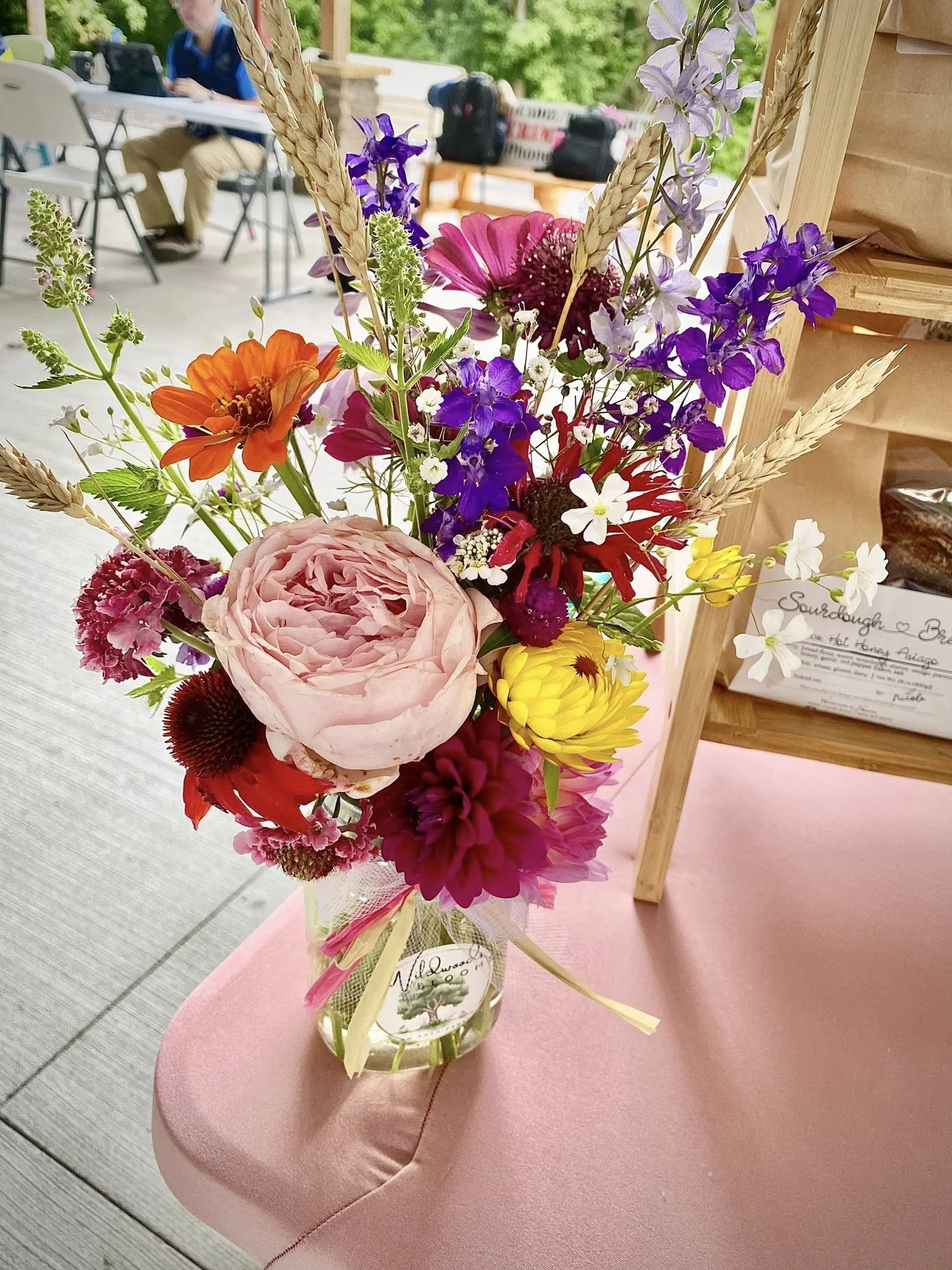 Colorful flower bouquet in a glass jar on a pink table, with a blurred outdoor background.
