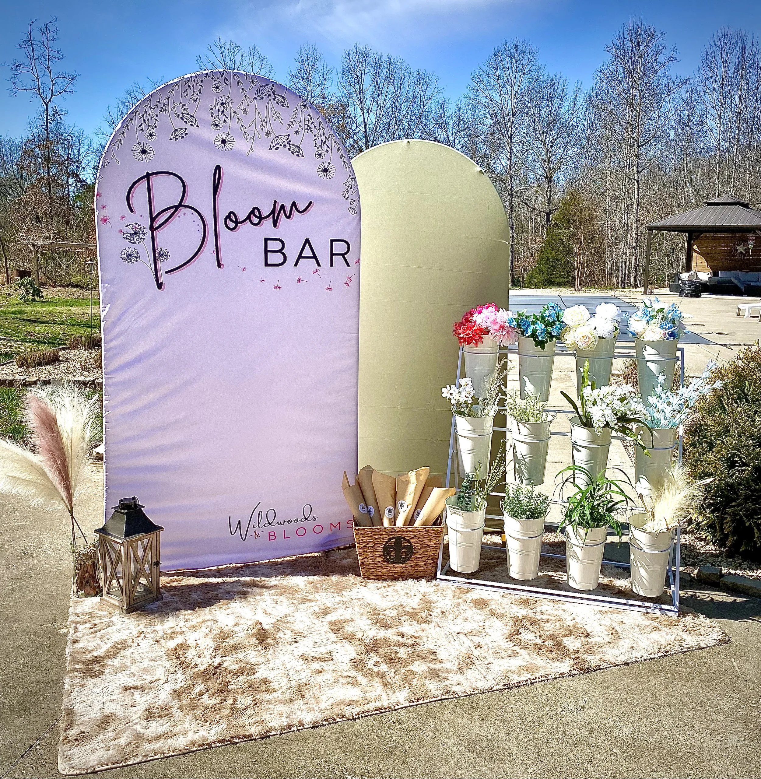 Decorative setup for Bloom Bar event with large pink and green panels, white flower buckets with colorful flowers, a lantern, and a basket of brown paper-wrapped items on a fluffy beige rug outdoors on a sunny day.