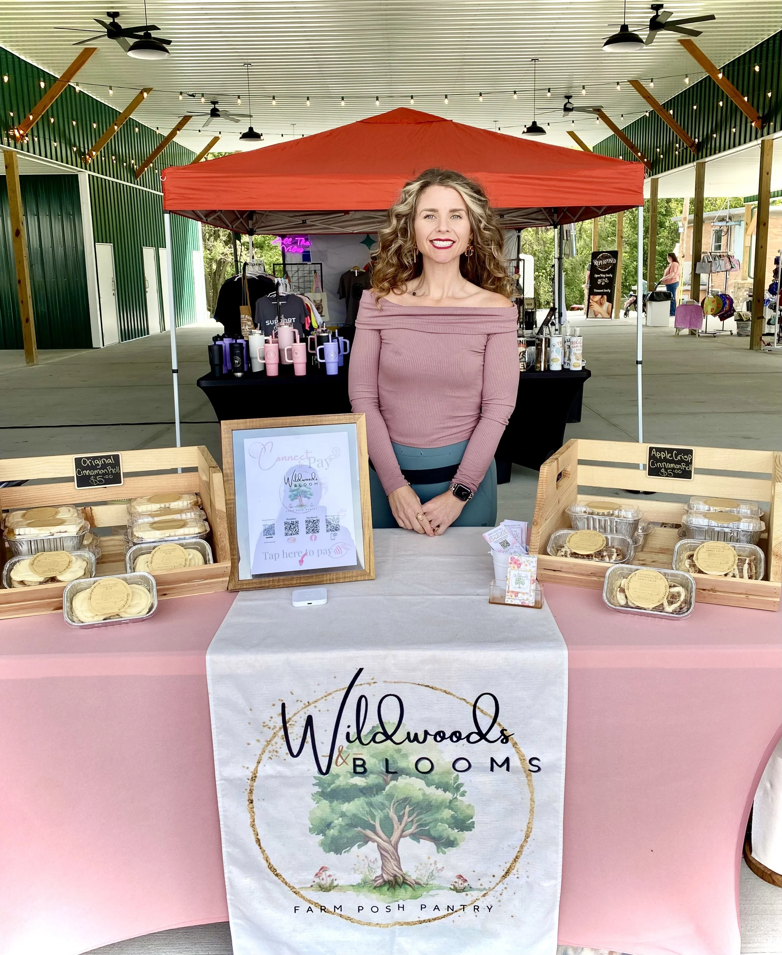 A woman standing behind a booth selling baked goods at an indoor or outdoor market. The booth has a sign that reads 'Wildwoods & Blooms Farm Posh Pantry' and various containers of baked items labeled 'Original Cinnamon Roll'. There are other products displayed behind her and a small framed sign on the table with a QR code for digital payment.
