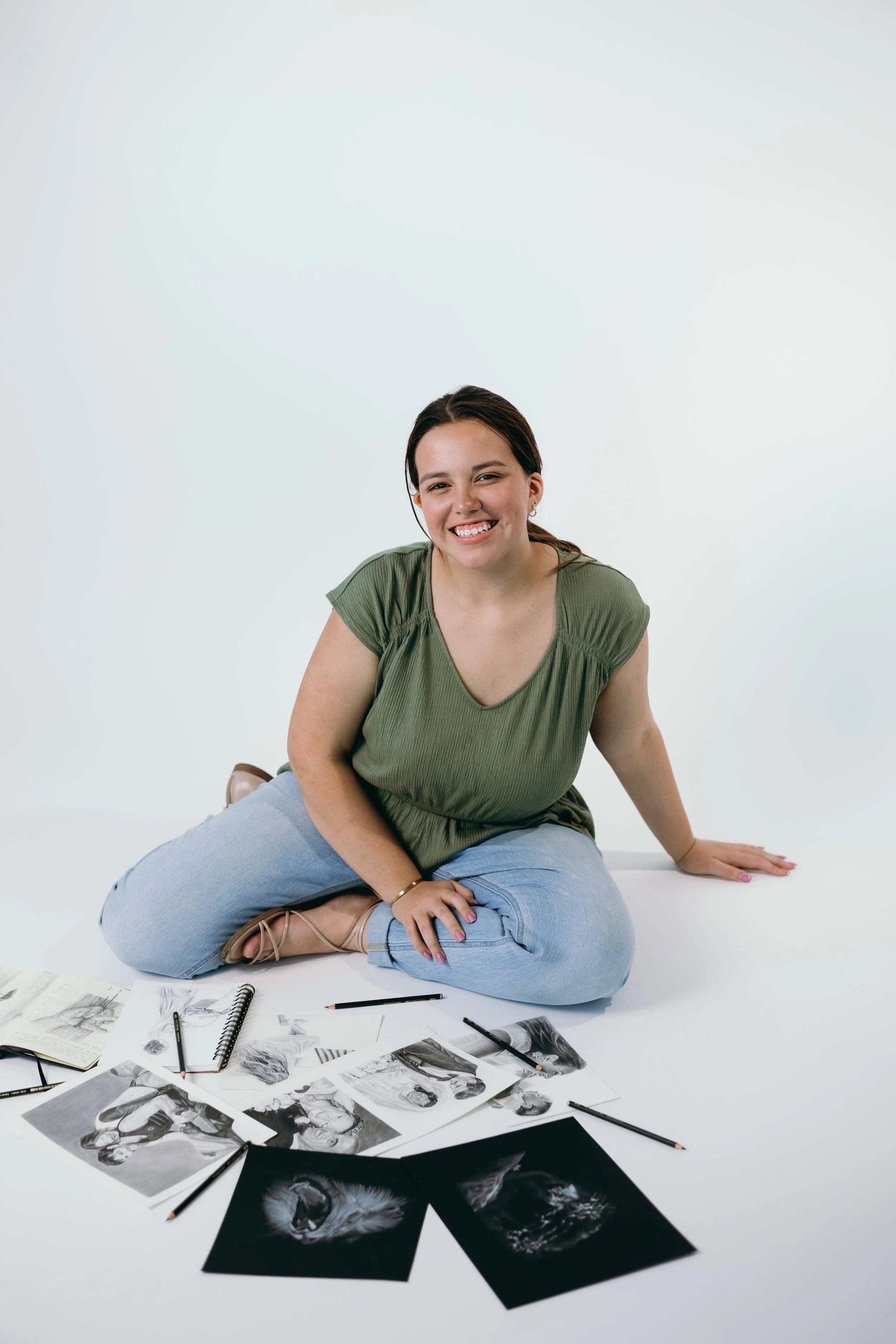 Smiling woman sitting on the floor with photographs of animals and sketches, surrounded by pens and notebooks, against a white background.