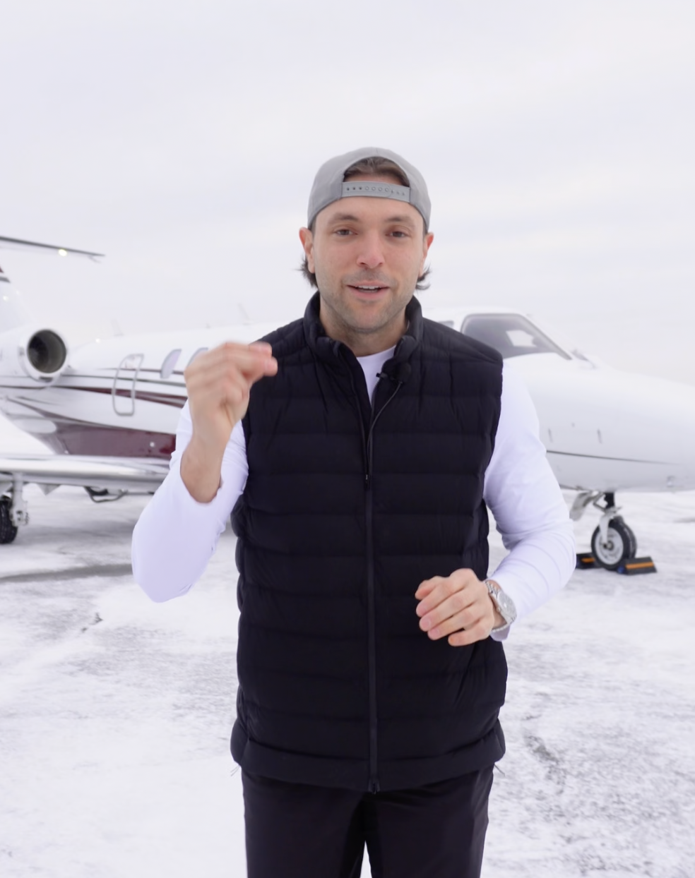 A man standing outdoors on a snowy tarmac with private planes in the background, wearing a gray cap, black vest, and white long-sleeve shirt, making a fist gesture.
