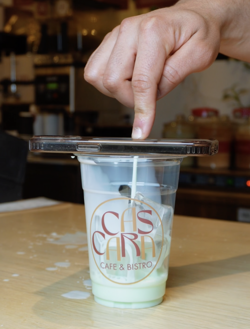 A hand with a finger pressing on a smartphone placed on top of a plastic cup with a café logo, liquid spilling around the cup, in a café or bistro setting.