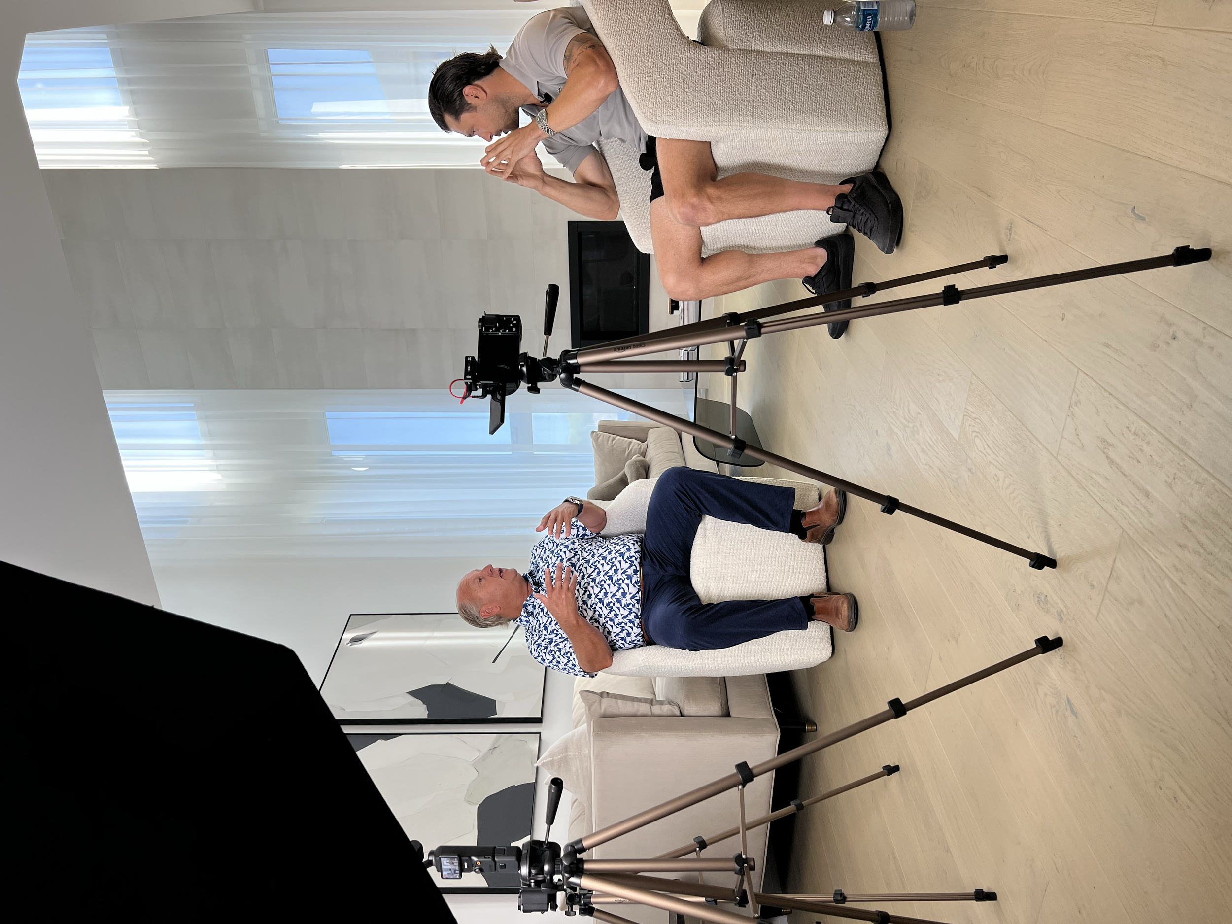 A man being interviewed or recorded sitting on a white armchair in a well-lit room with modern decor. A woman is seated nearby, possibly part of the interview team, with film or camera equipment set up and focused on the man.