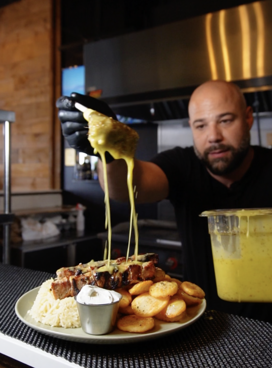 A man is pouring melted cheese onto a plate of barbecue ribs, potato chips, rice, and a small cup of whipped cream in a restaurant kitchen.