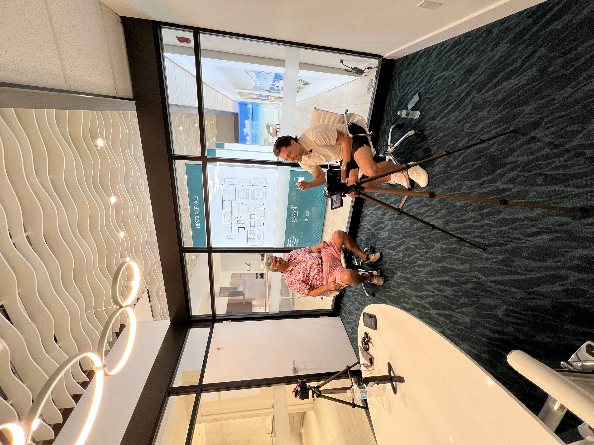 Two people engaged in a discussion or interview in a modern office with a large glass partition and a screen displaying a building floor plan. One person is seated while the other is standing, both in casual attire. Equipment like a tripod and camera