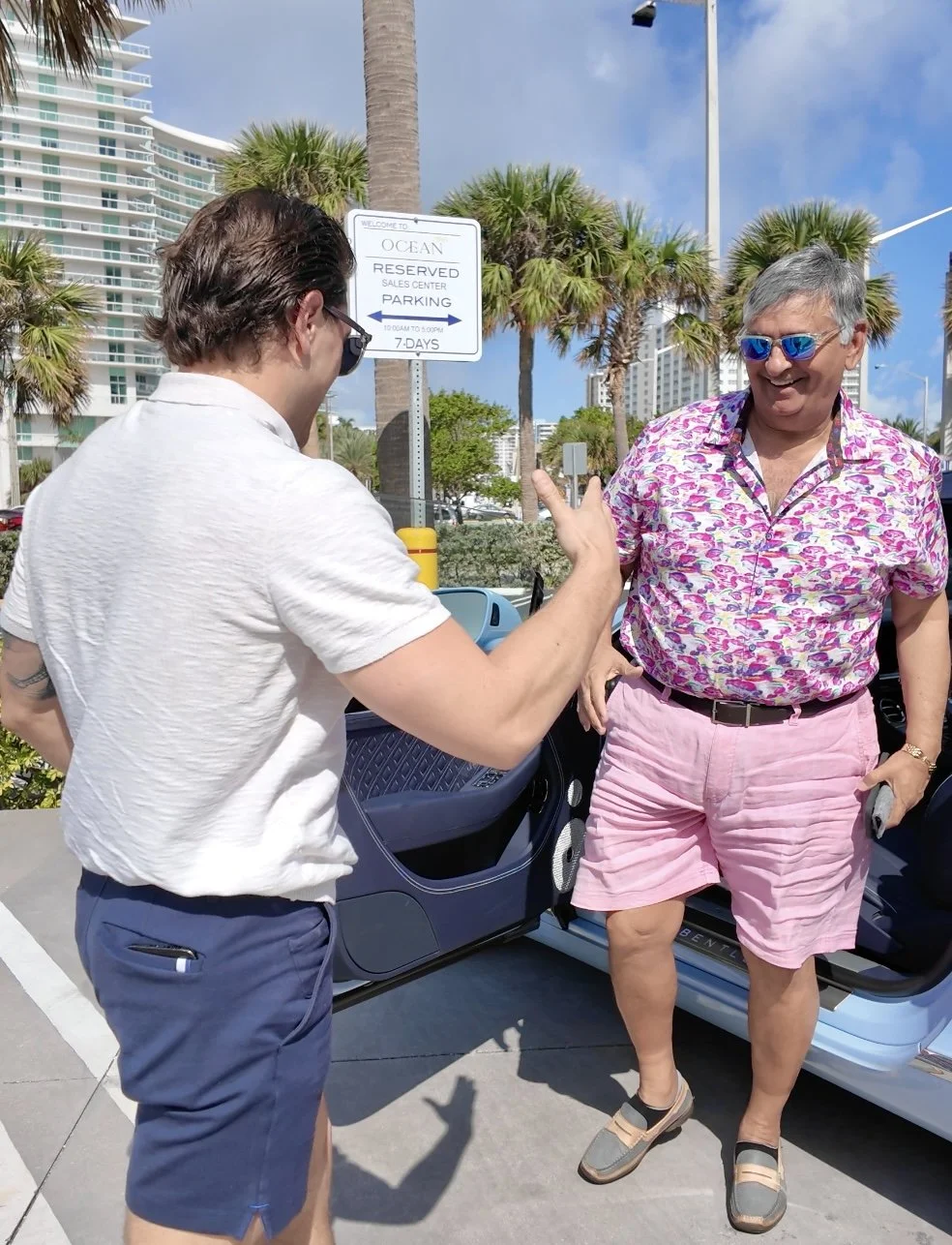 Two men standing outside near a parking lot, having a friendly conversation. The man on the left is wearing a white shirt and navy shorts, and the man on the right is wearing a colorful pink shirt and pink shorts with sunglasses. There are palm trees