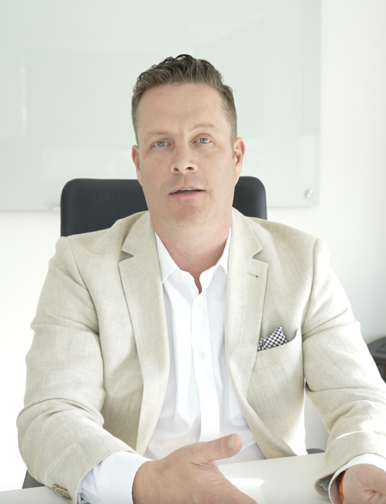 A man in a beige blazer and white shirt sitting at a desk in an office, looking at the camera.