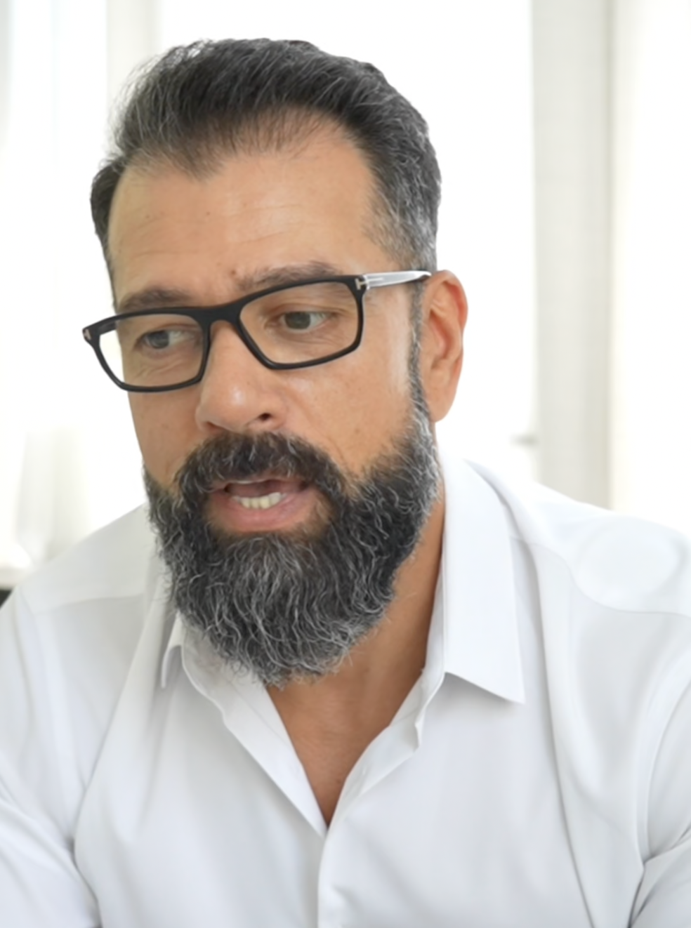 A man with glasses, a gray beard, and a white dress shirt is speaking or listening with a serious expression, in a bright room.
