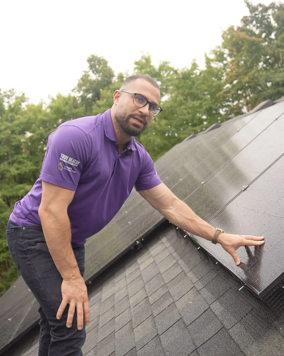 Man in glasses and purple polo shirt inspecting solar panels on a sloped roof during daytime with trees in the background.