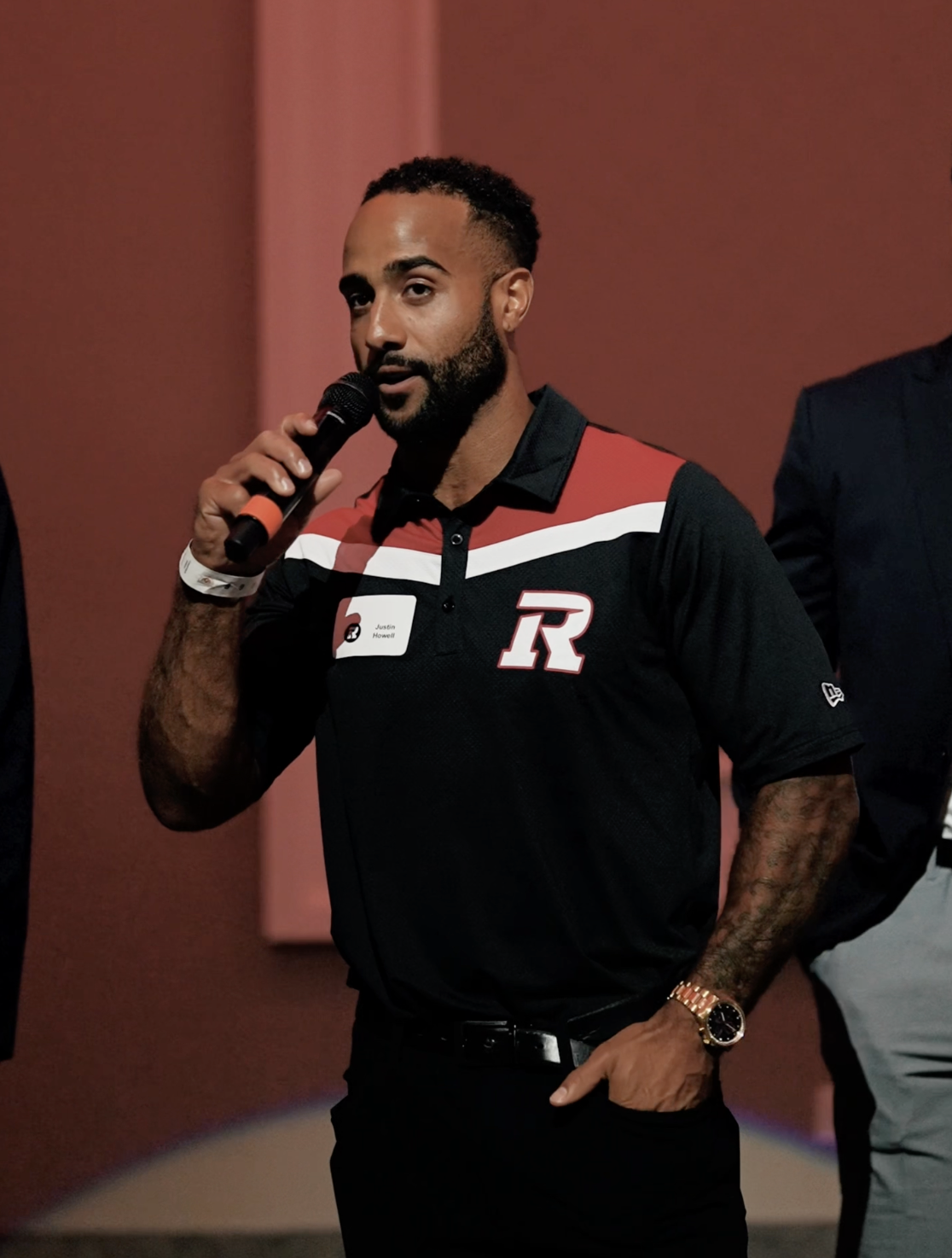 Man with a beard wearing a black and red polo shirt holding a microphone, with a stage background.