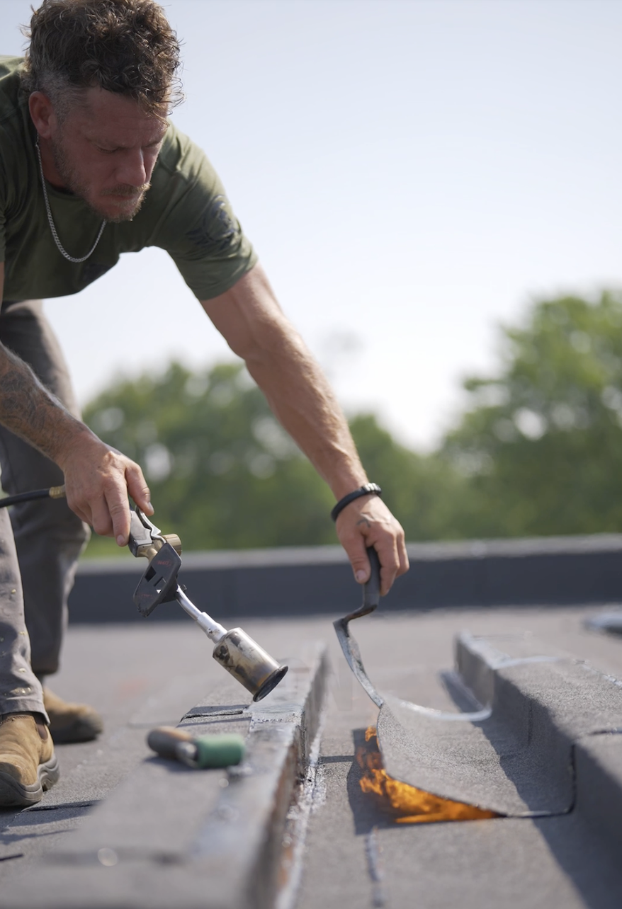 Man working on a rooftop, using a blowtorch and a crowbar to repair or install roofing materials, with trees in the background.