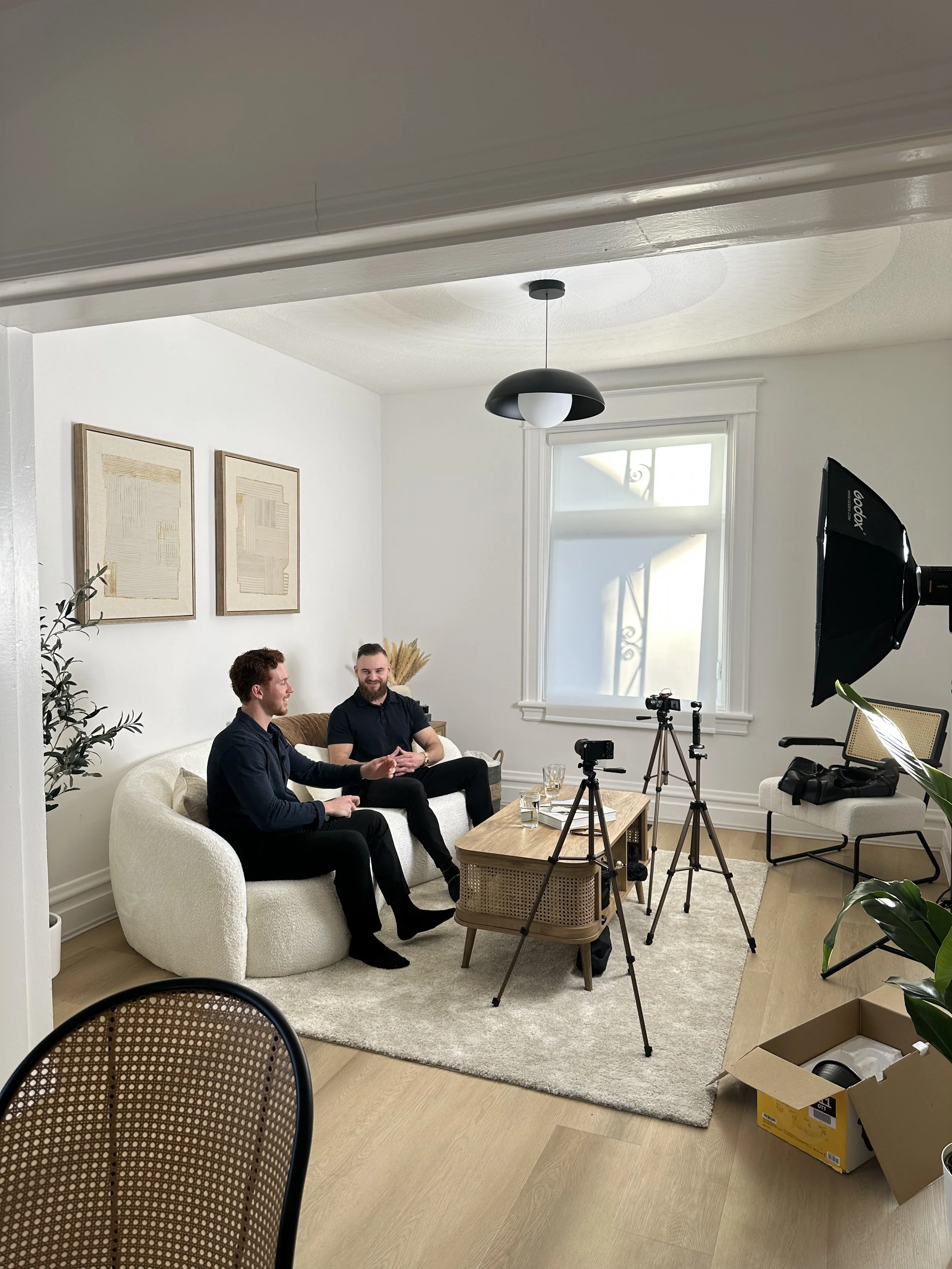 Two men sitting on a cream-colored sofa in a bright living room, recording a video or podcast with camera equipment and a softbox light set up nearby.