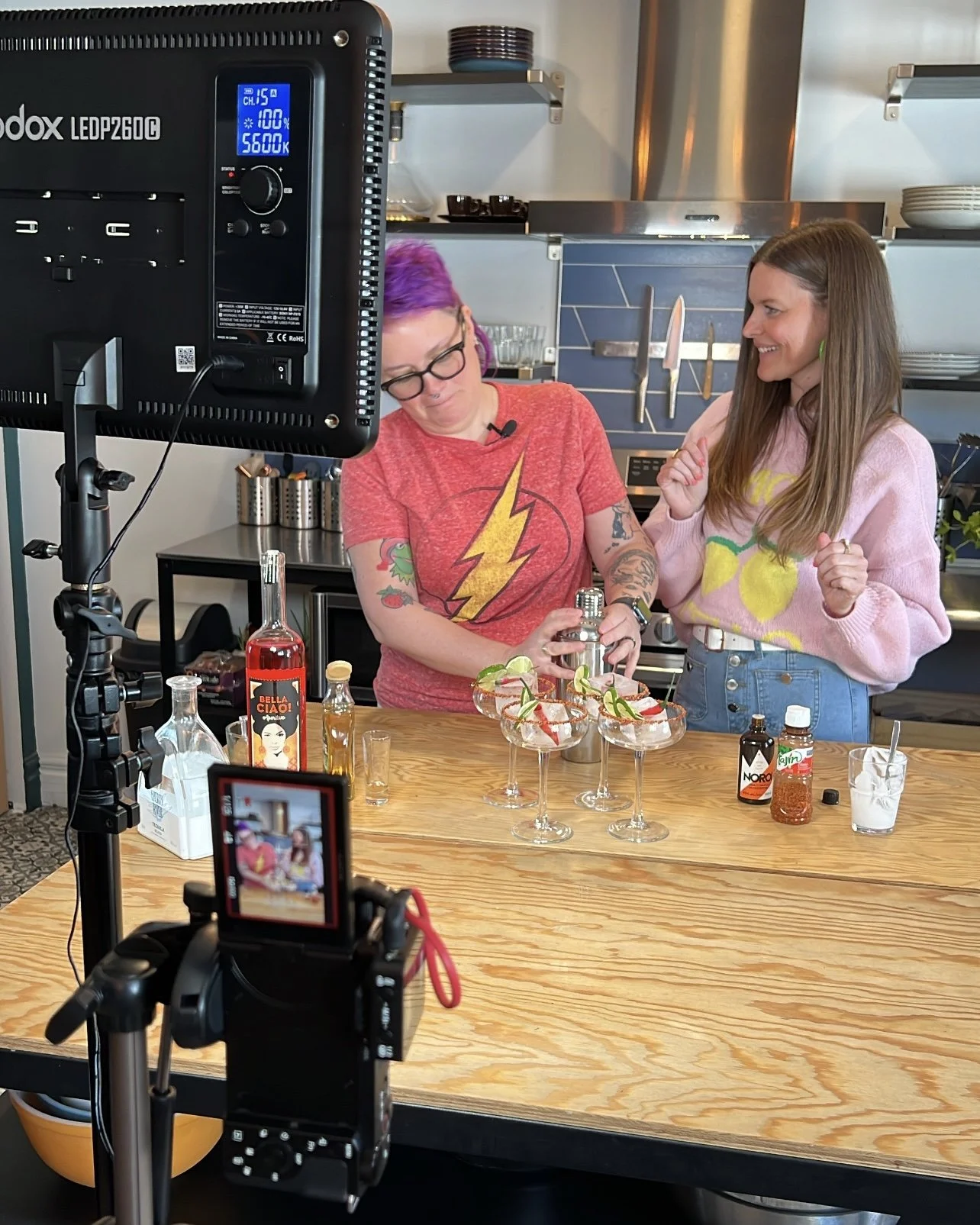 Two women preparing cocktails in a kitchen studio. One woman with purple hair and glasses wearing a red t-shirt with a lightning bolt, and another woman with long brown hair wearing a pink sweater with a lemon print, smiling and talking. Various bott