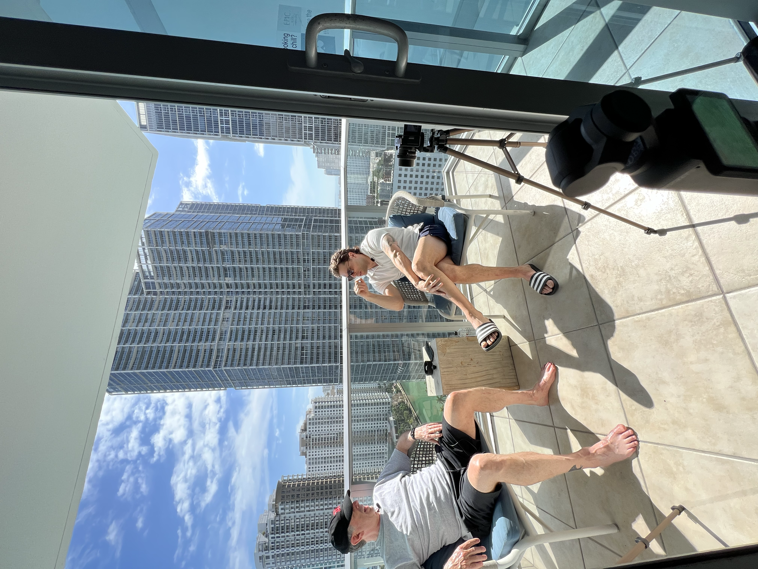 A man sitting on a balcony talking to a woman, with tall skyscrapers in the background, a camera on a tripod, and telescope. The man is barefoot, wearing black shorts, a gray t-shirt, and a black cap. The woman is wearing slides, shorts, and a top, a