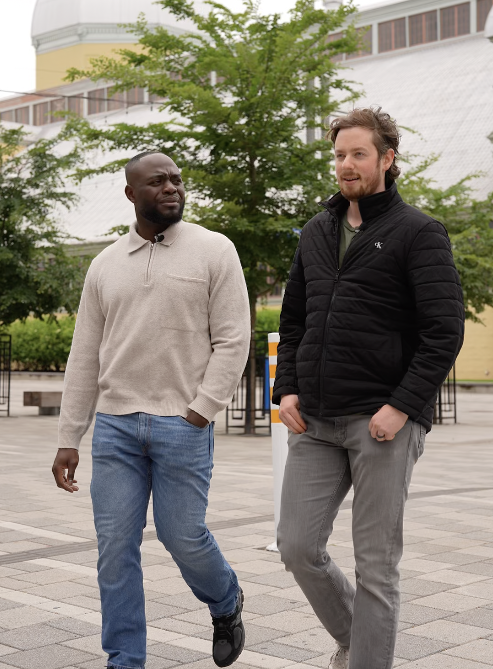 Two men walking and talking outdoors on a paved area with trees and a building in the background.