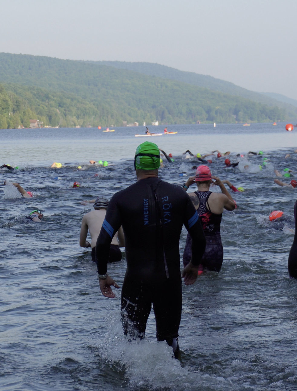 Swimmers in wetsuits and swim caps entering a lake for a race, with a backdrop of hills and kayakers on the water.