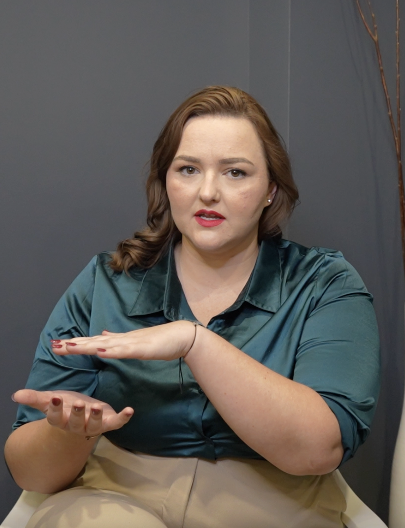 A woman with shoulder-length brown hair, wearing a teal satin blouse and beige pants, is sitting in front of a gray wall and gesturing with her hands as if explaining something.