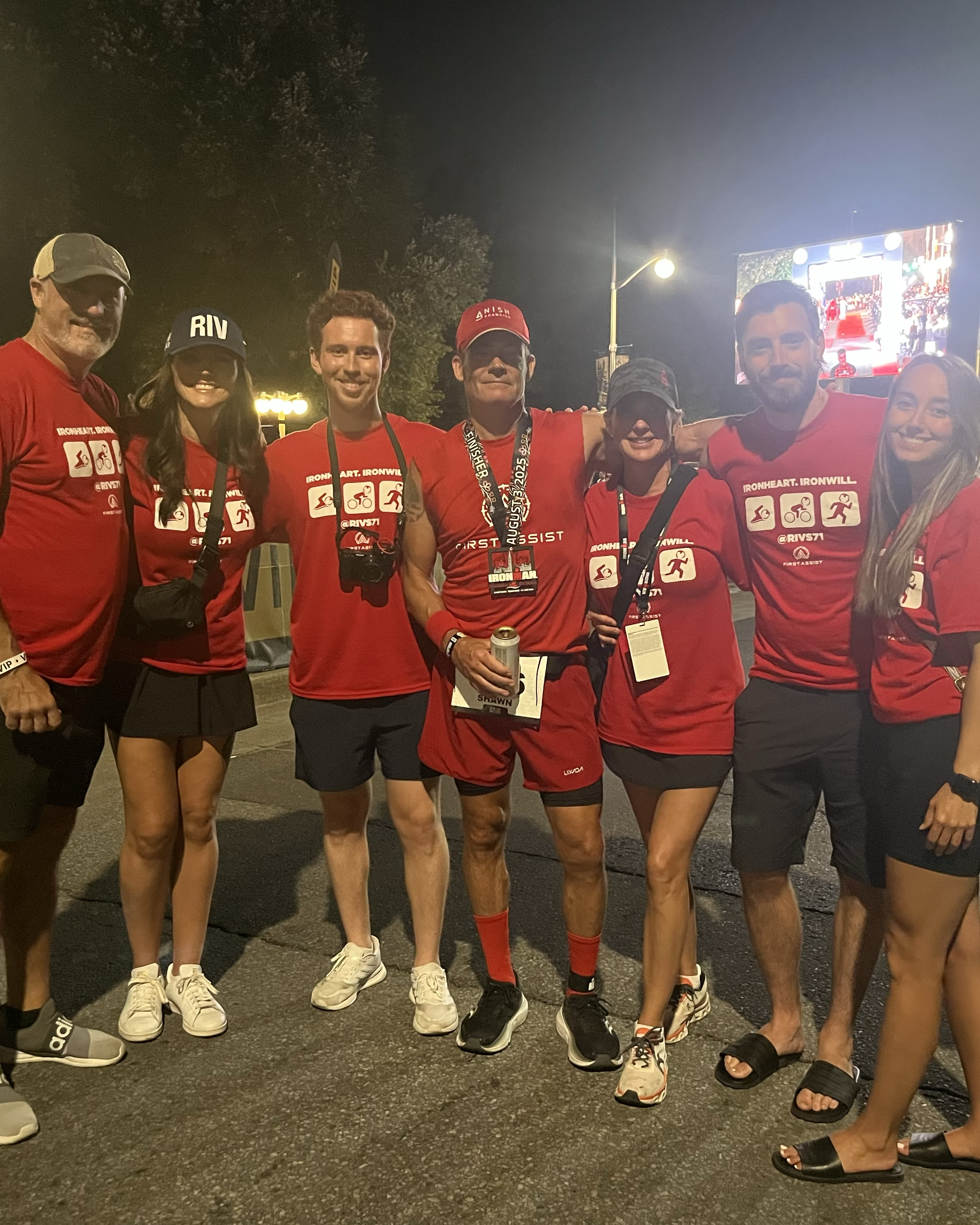 Group of seven people posing at night, all wearing red shirts with 'IronHeart IronWill' and icons of sports activities, some holding drinks, with a large screen in the background displaying a similar outdoor event.
