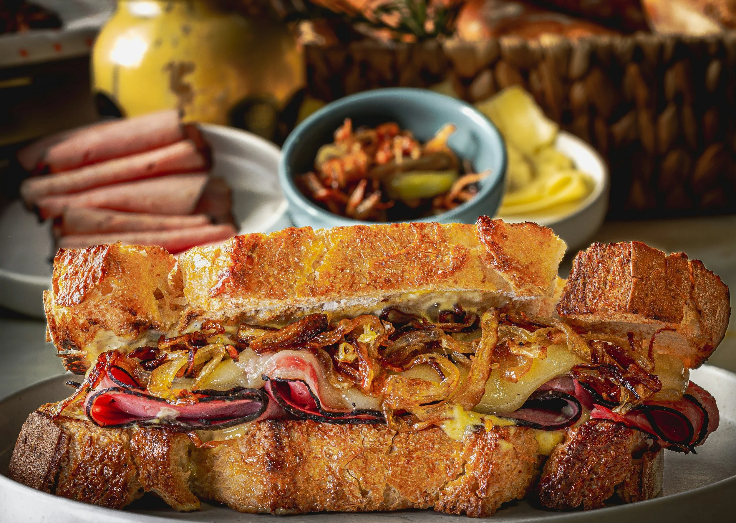 A savory French dip sandwich with roast beef, melted cheese, caramelized onions, and toasted bread, served with side dishes including sliced ham, pickles, and a small bowl of pickled vegetables.
