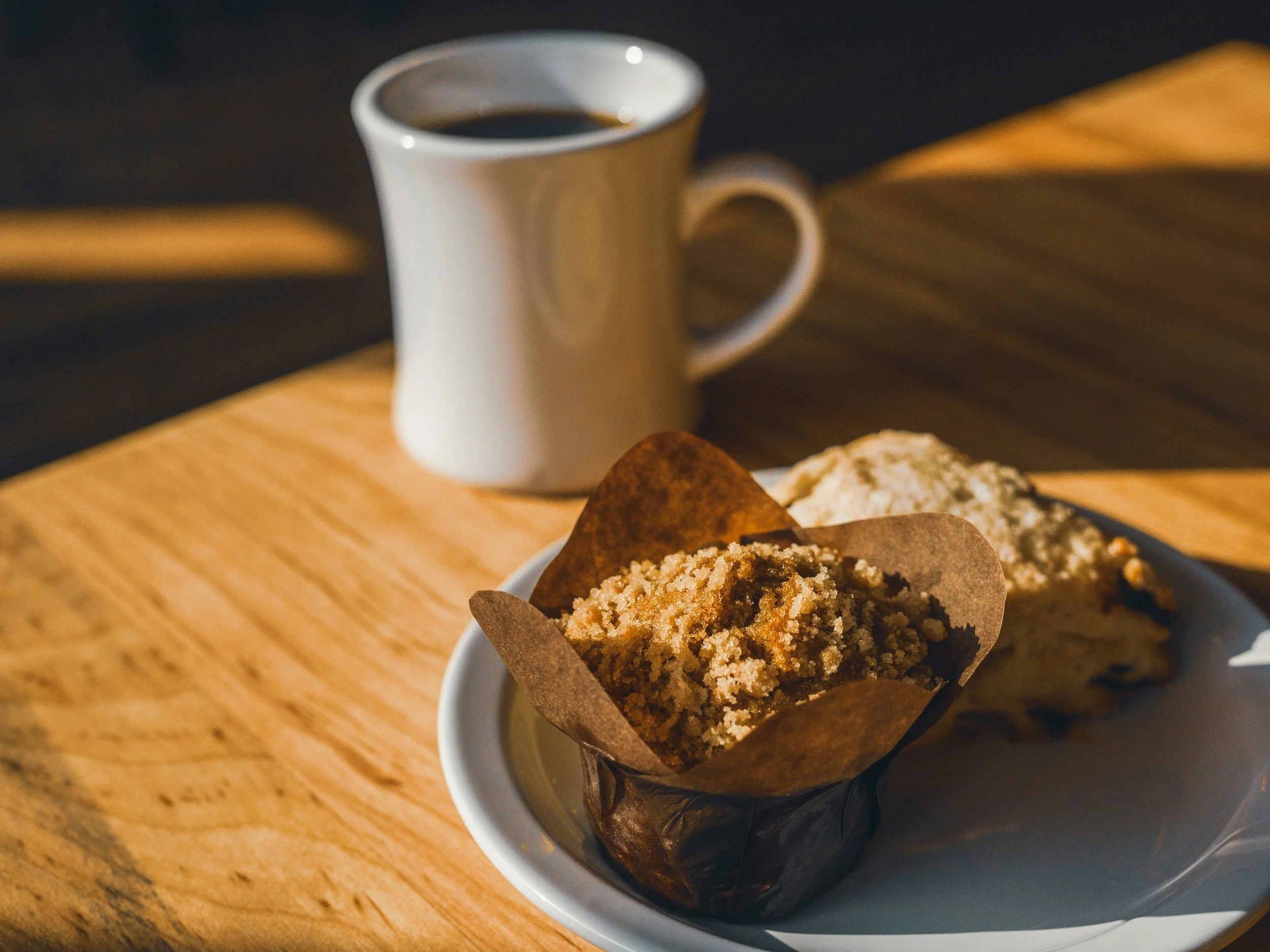 A white plate with two baked goods, one topped with crumble and the other possibly a muffin or scone, on a wooden table with a coffee mug filled with black coffee in the background.