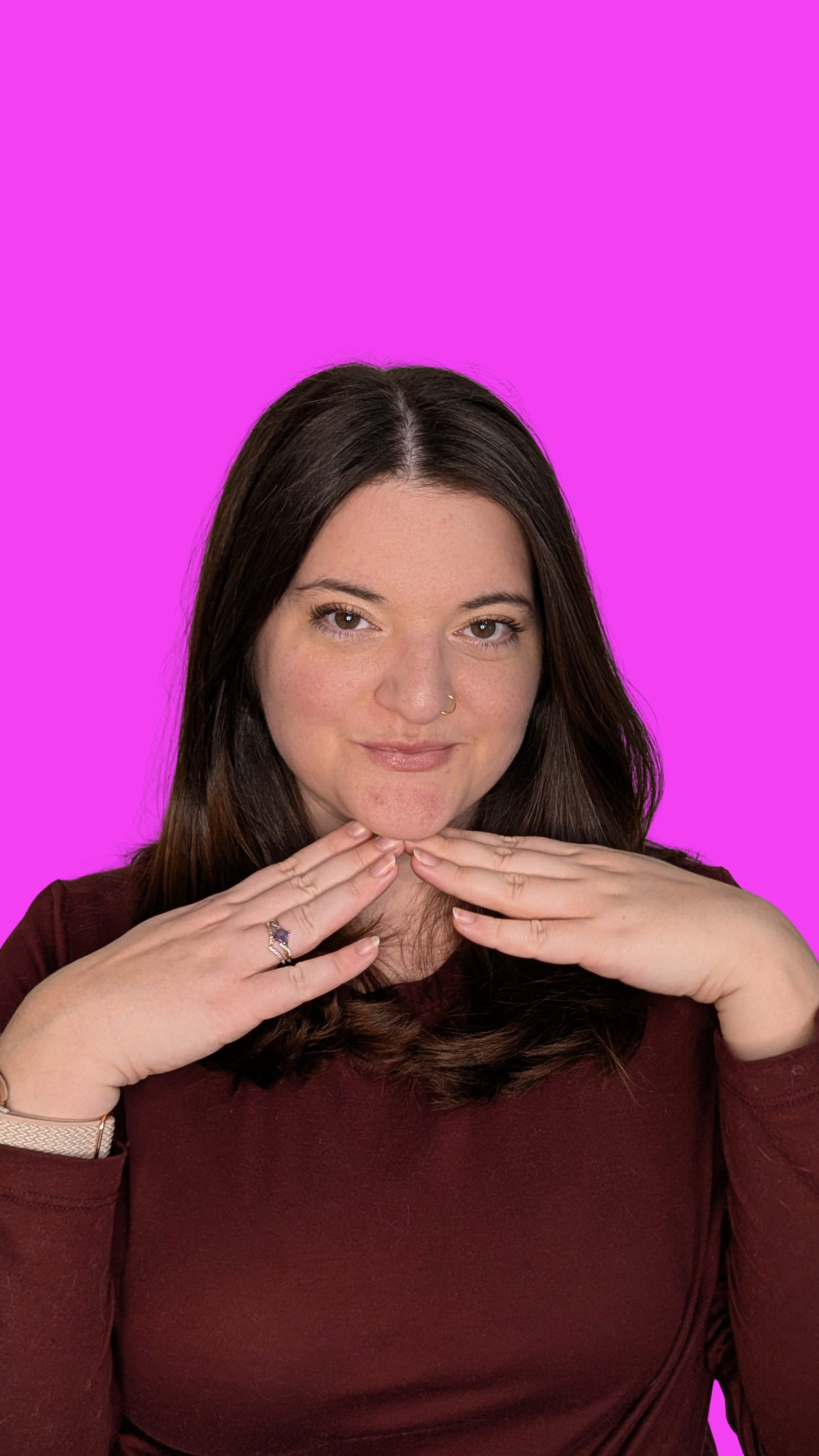 A woman with brown hair, wearing a maroon shirt, making a playful pose with her hands under her chin, against a bright pink background.